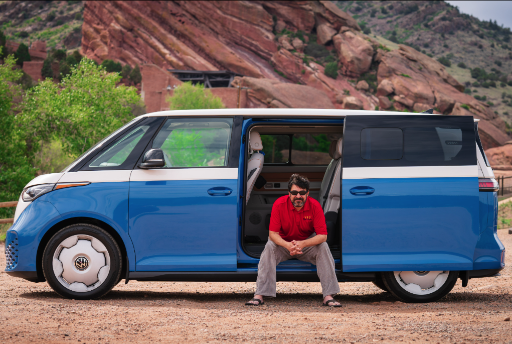 Man with sunglasses and a red shirt sitting on the side step of a blue and white Volkswagen electric van parked outdoors in a desert landscape with rocky hills and some green trees.