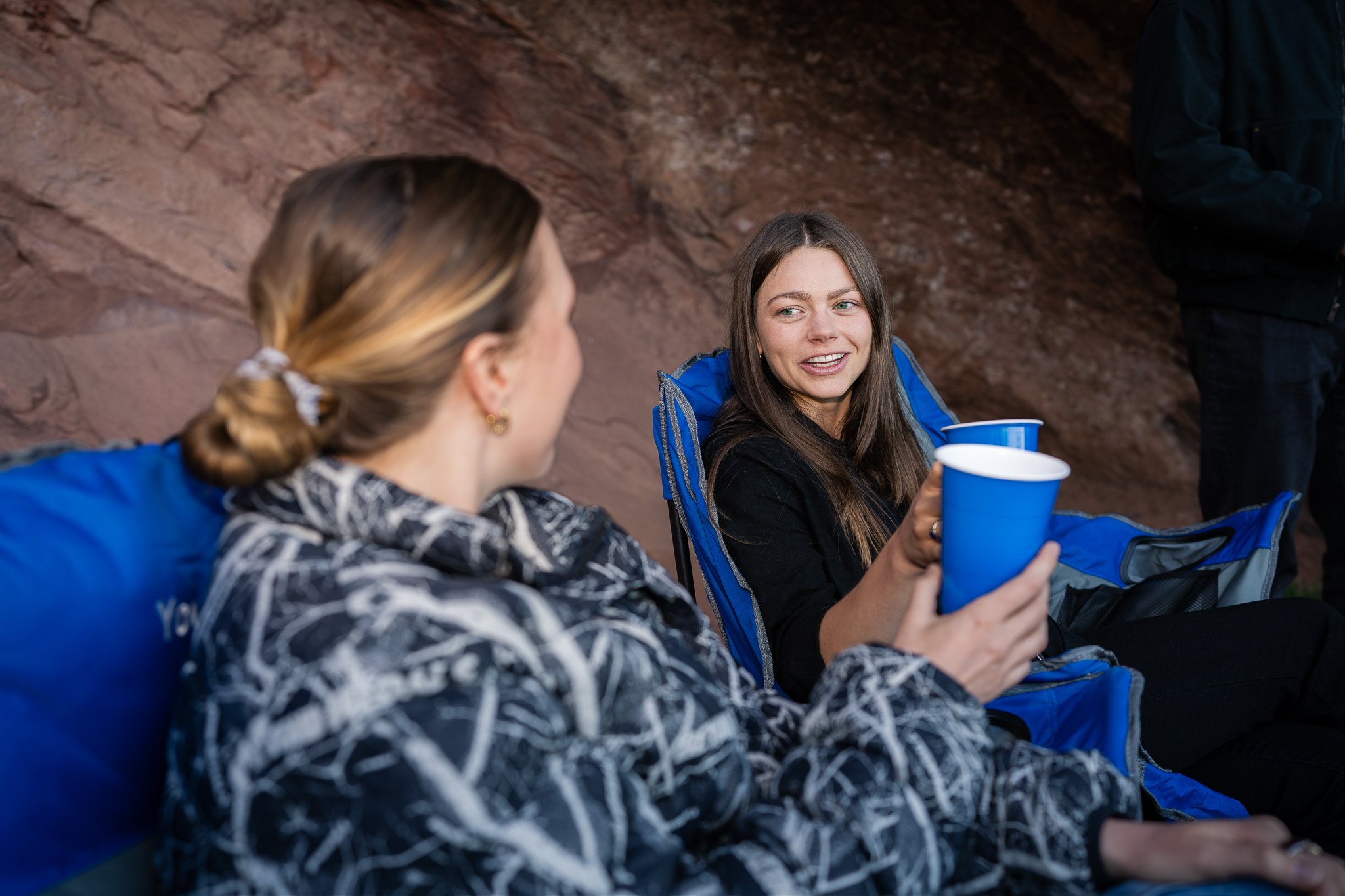 Two women sitting next to eachother at the tailgate in the Red Rocks upper parking lot, holding blue cups and enjoying custom mixers while hanging out with friends before the event starts.