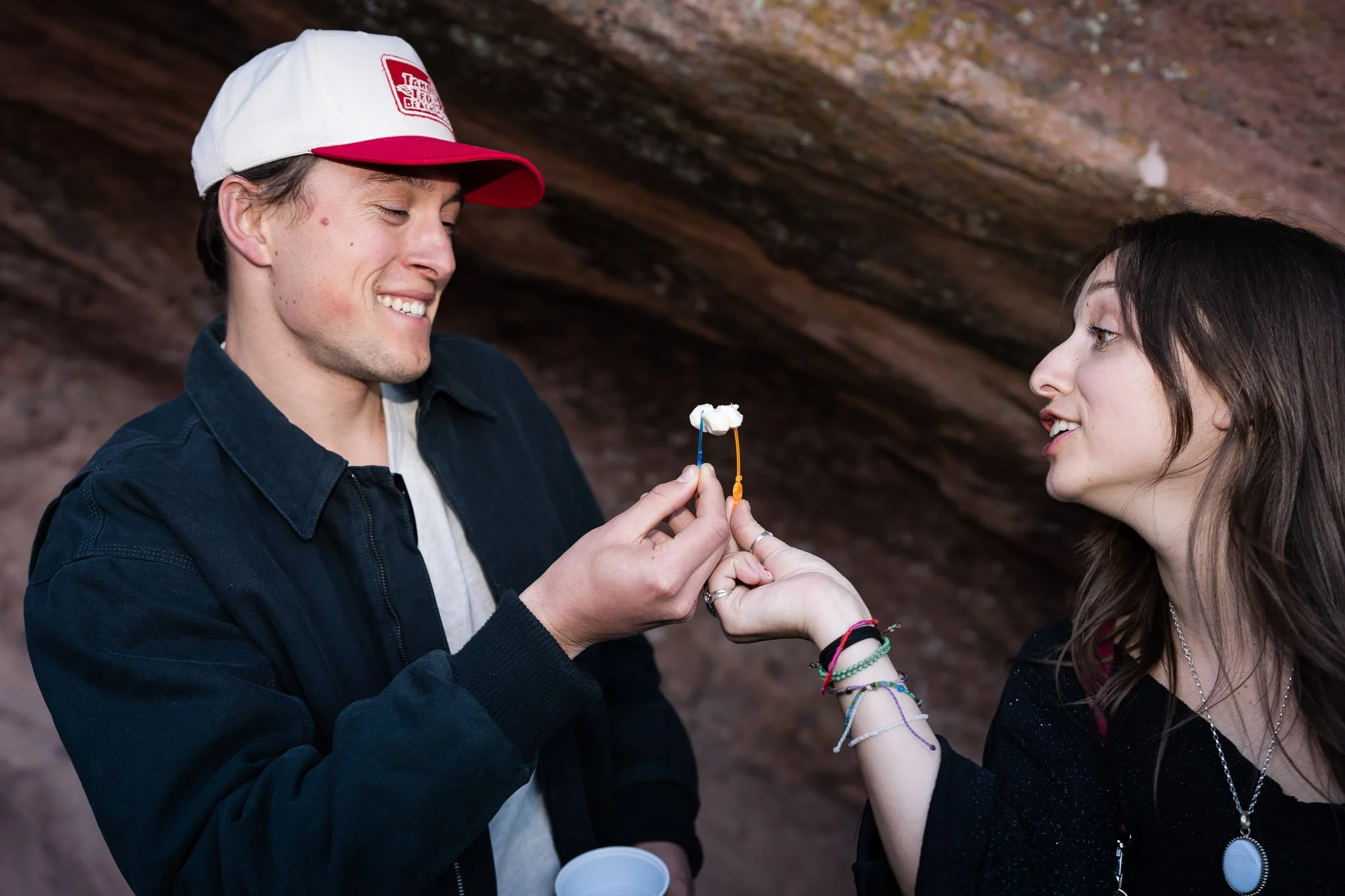 A young man with a hat and a young woman with bracelets cheers-ing their tailgate charcuterie board cheese before the show at Red Rocks.