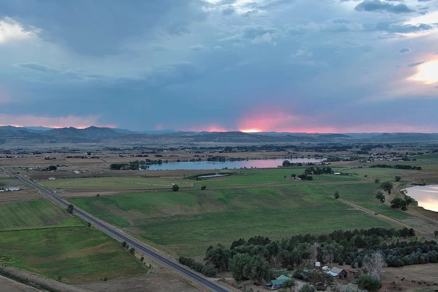 A scenic view of flat farmland with patches of green fields, small lakes, and scattered trees, with mountains on the horizon and a sky filled with dark and pink-tinged clouds at sunset.