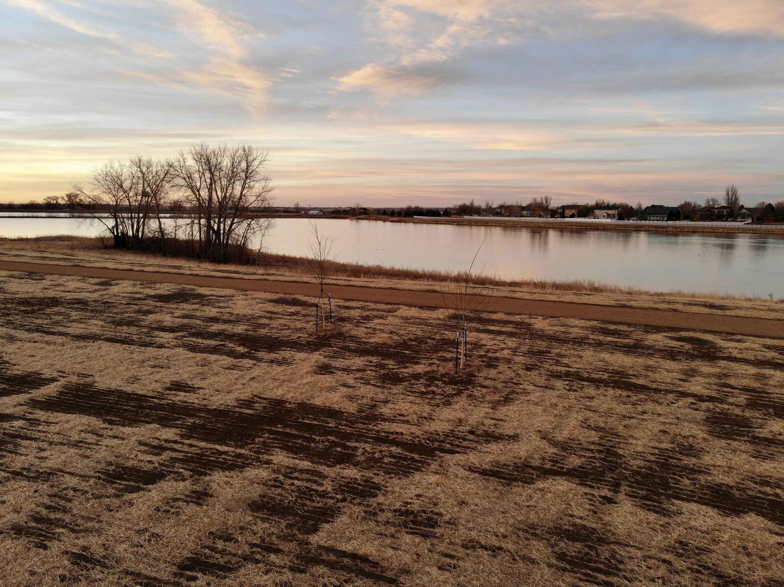 A serene landscape of a lake at sunset with a partly cloudy sky. On the foreground, there is a dirt path and leafless trees. In the background, some residential houses are visible along the far shoreline.