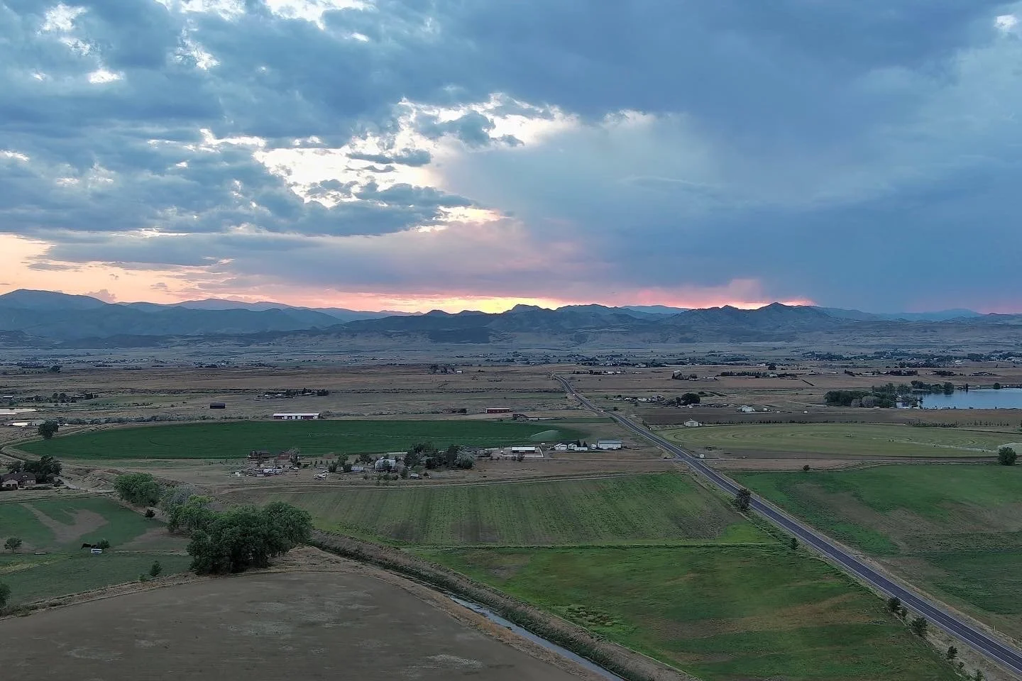 A scenic landscape at sunset showing green farmland, a distant mountain range, a road cutting through the fields, and a partly cloudy sky with sunlight peeking through the clouds.