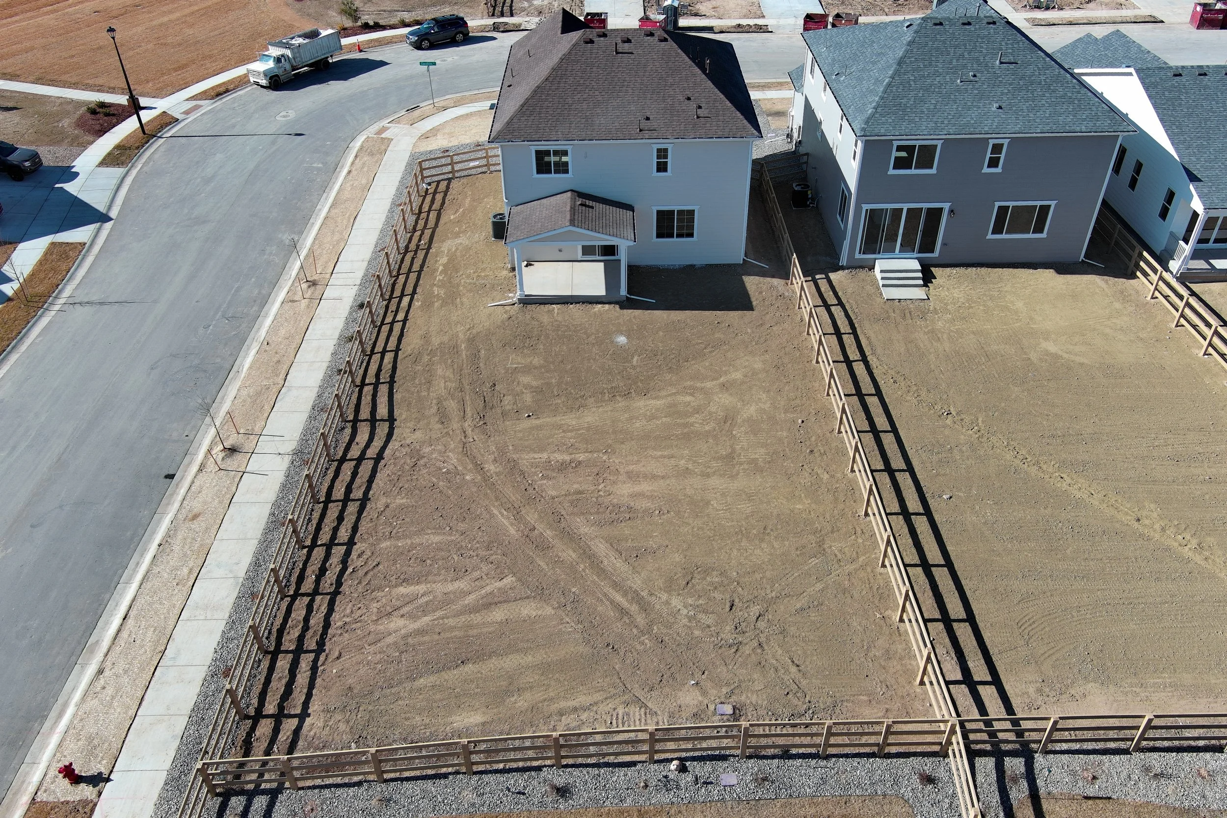 Aerial view of a residential area showing two houses with fenced yards, a street with parked cars, and a lot of bare dirt in the backyard of one house.