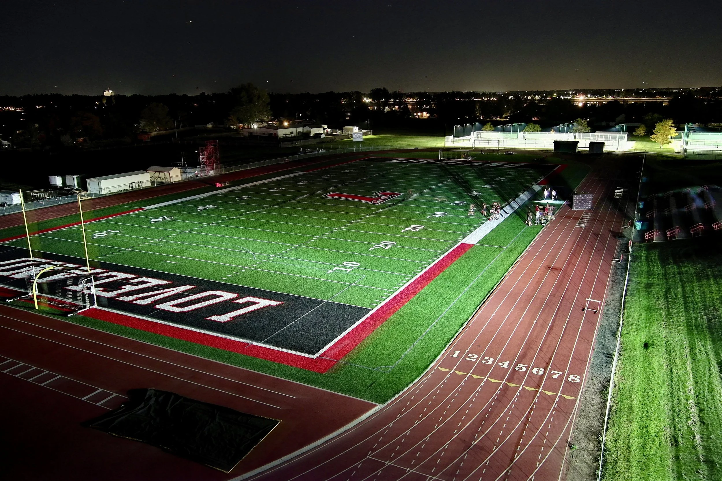 Night view of a football stadium with brightly lit field and track, some people gathered along the side, and a city skyline with lights in the background.