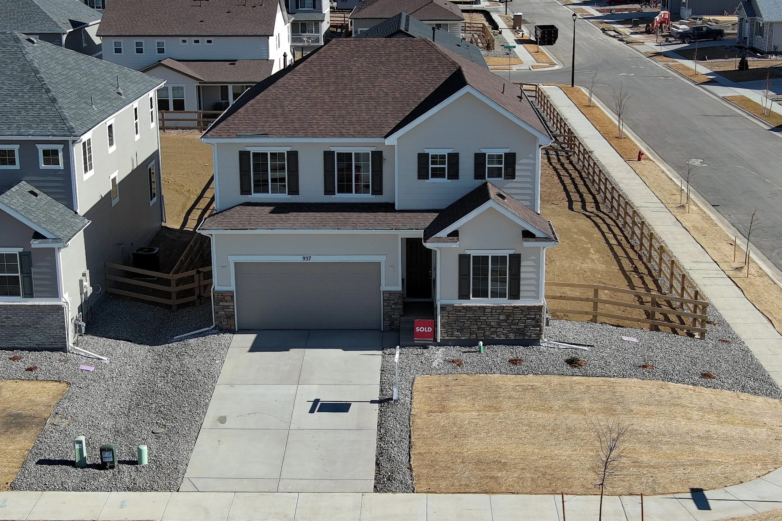A two-story house located in a neighborhood, with a brown roof, white exterior, and black window shutters. The driveway and sidewalk are visible, with a "Sold" sign in front of the house. Empty plots with young trees and fences surround the house.