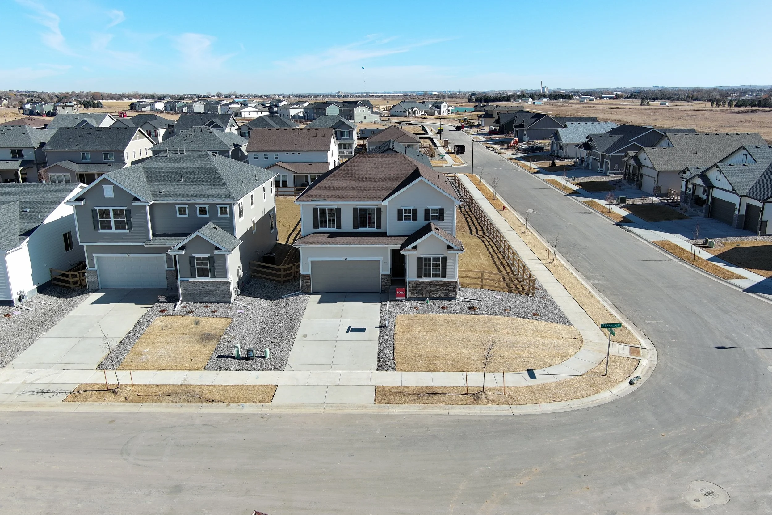 Aerial view of new suburban neighborhood with modern houses, paved streets, and open fields in the background on a clear day.