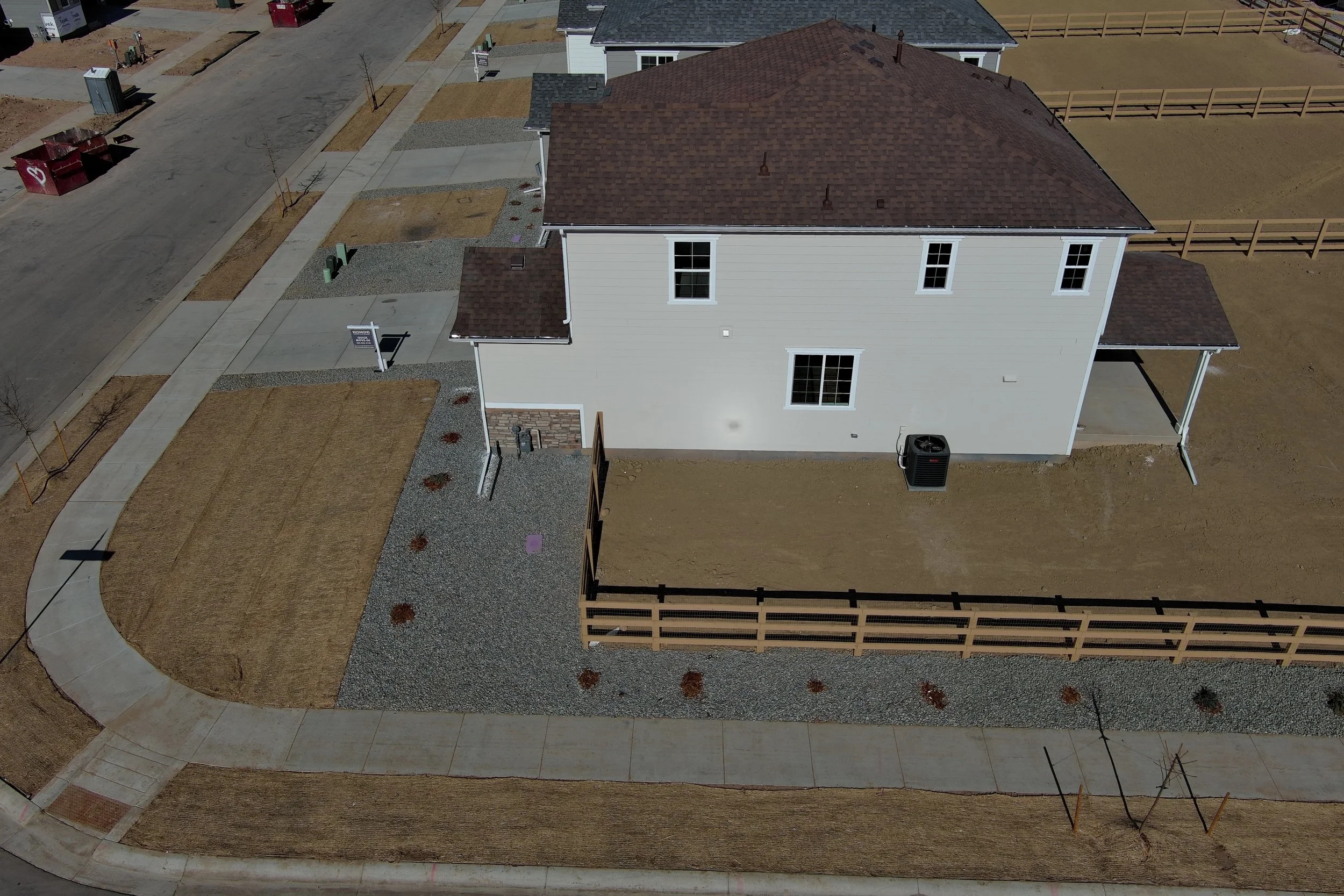 Aerial view of a new residential neighborhood under construction showing a white house with a brown roof, concrete sidewalks, and neighboring areas with dirt and landscaping.