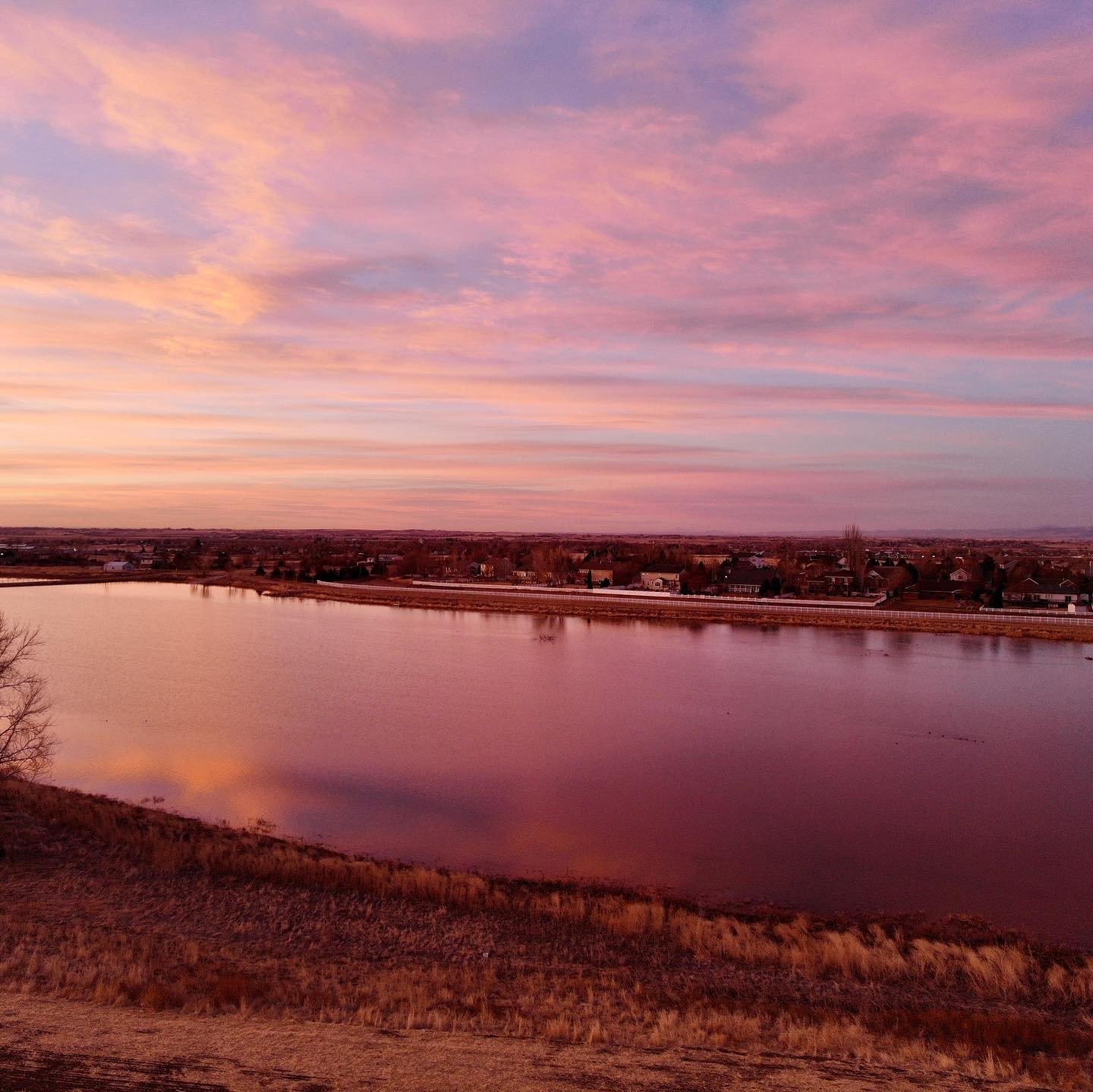 A peaceful river reflecting a colorful sunset sky with pink, purple, and orange clouds, and a distant town along the shoreline.