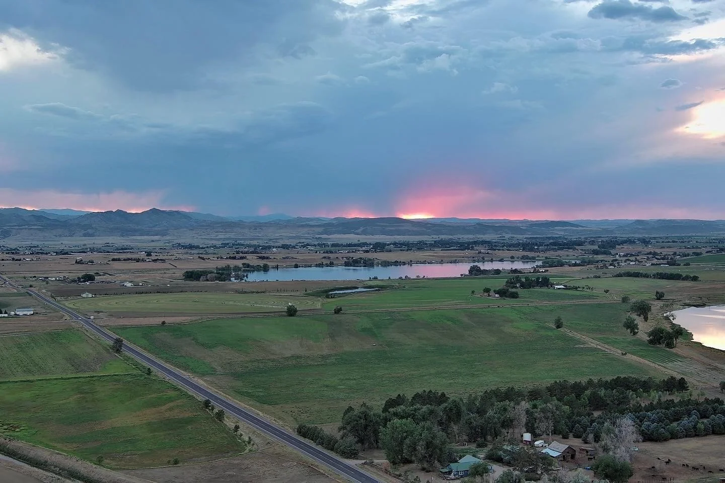A landscape with farmland, lakes, and distant mountains during sunset or sunrise, with dark clouds and pinkish glow near the horizon.