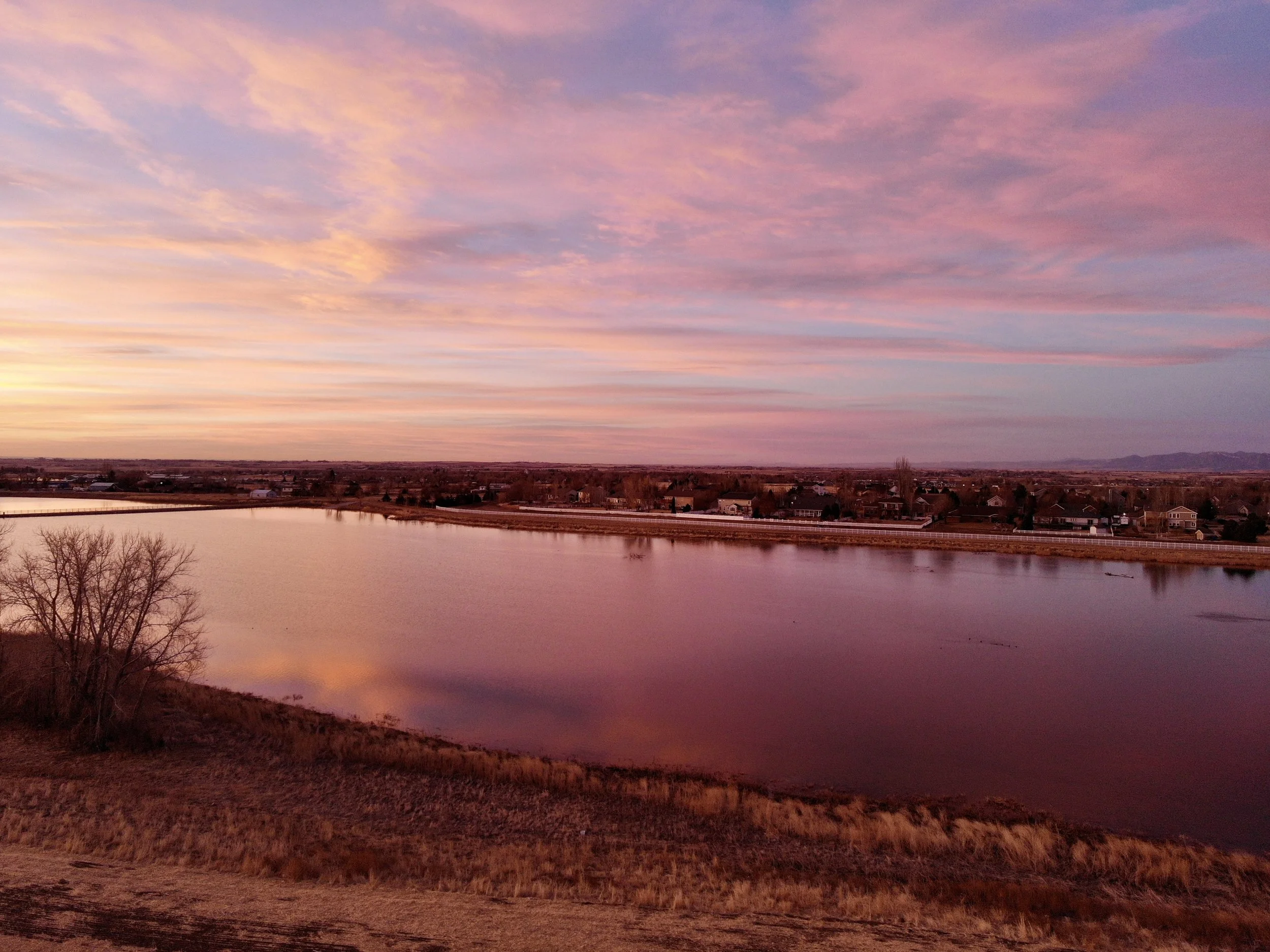 A scenic view of a large body of water reflecting a colorful sunset sky with shades of pink, purple, and orange. The horizon shows a small town or residential area with houses and trees, with mountains in the distance.
