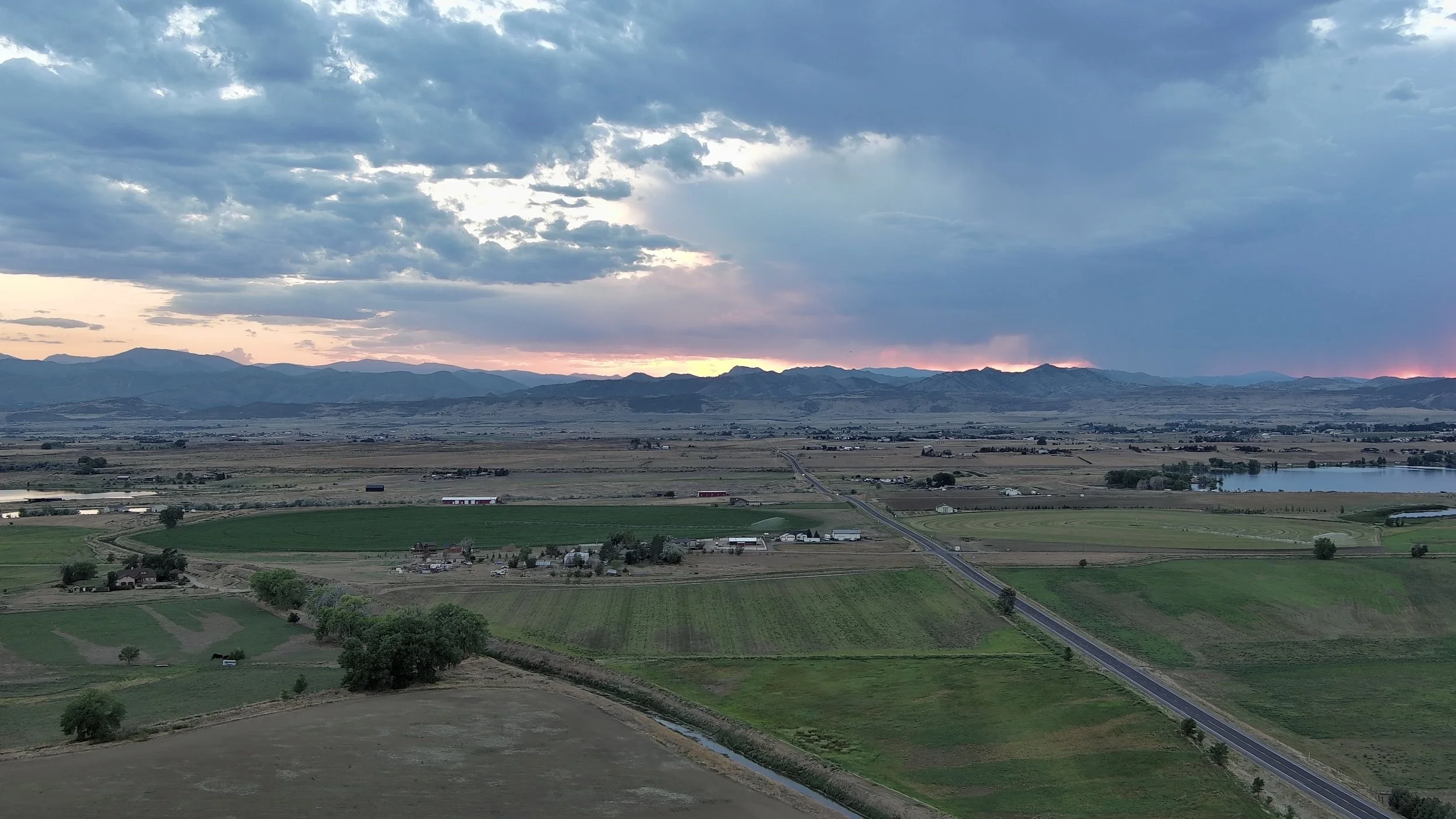 A wide landscape view of farmland and mountains at dusk, with a partly cloudy sky and a faint sunset light.