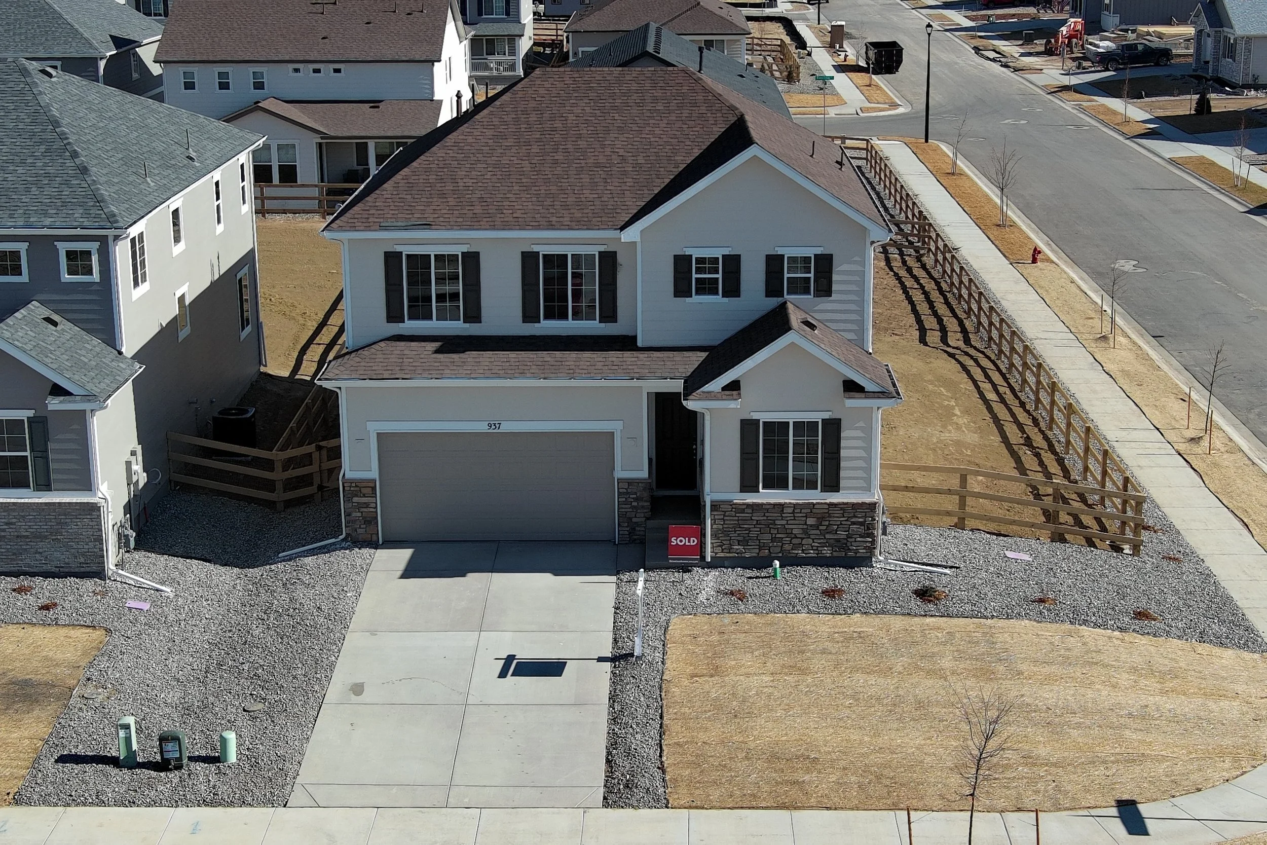 A newly constructed two-story house with a brown shingle roof, white siding, and black shutters, located in a suburban neighborhood. The house has a front yard with a concrete driveway leading to an attached garage, with a 