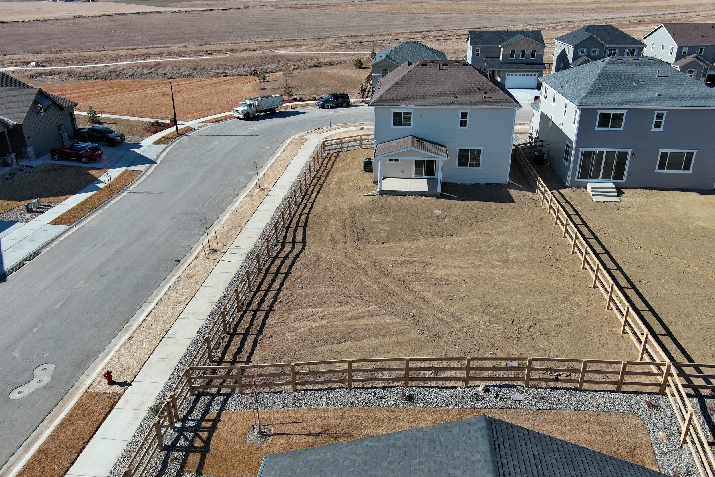 Aerial view of a suburban neighborhood with newly built houses, a curved street, parked cars, and an empty yard with a fence.
