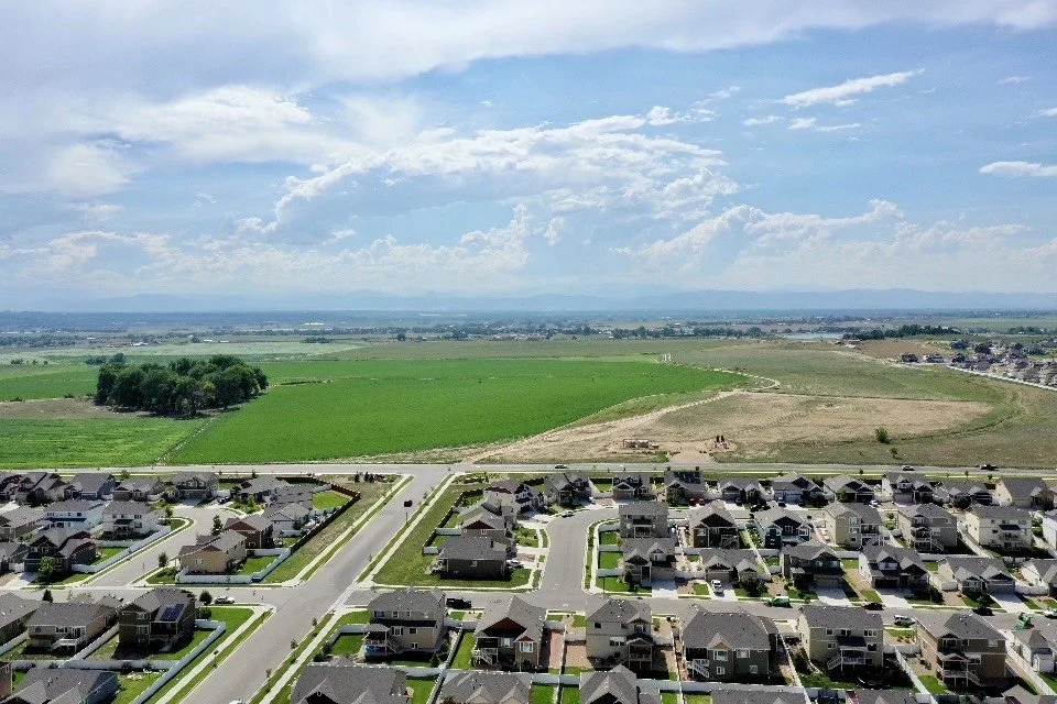 A suburban neighborhood with rows of houses, green lawns, and streets, with open farmland and a blue sky with clouds in the background.