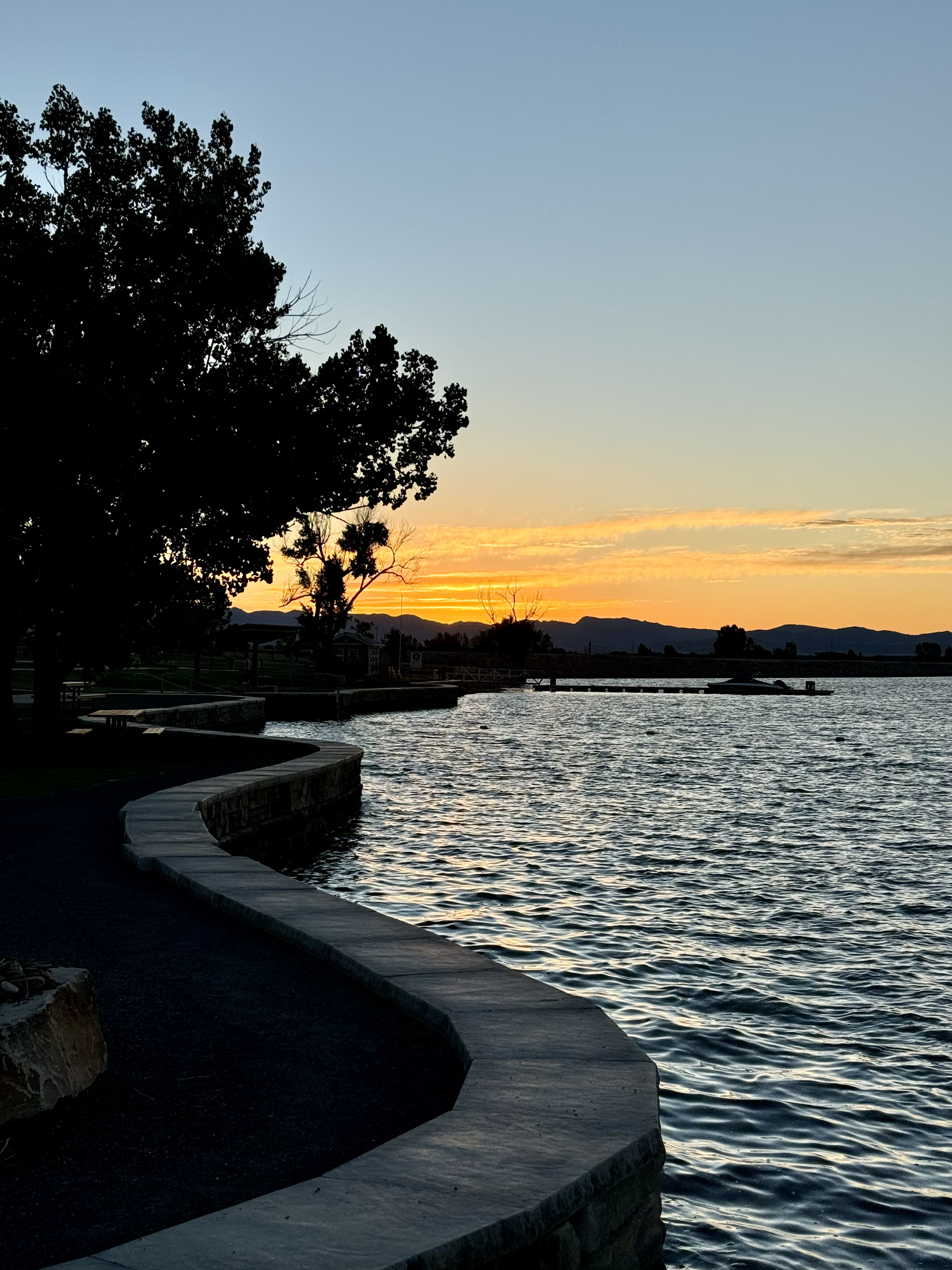 Sunset over a lake with silhouettes of trees and mountains in the background, calm water reflecting the sky, walkways along the shore.