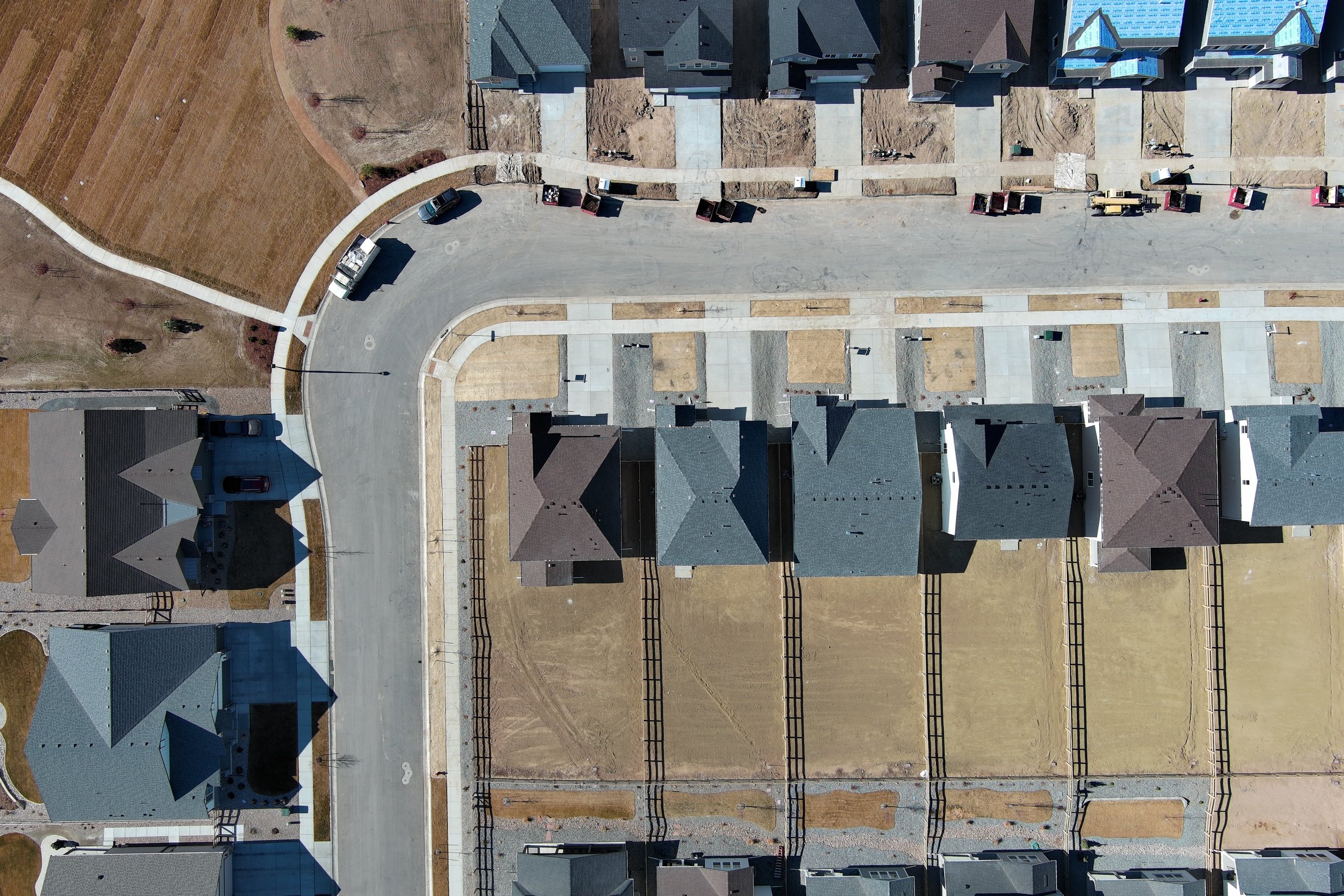 Aerial view of a residential neighborhood with houses, sidewalks, and an empty street under construction, with construction machinery and trucks.
