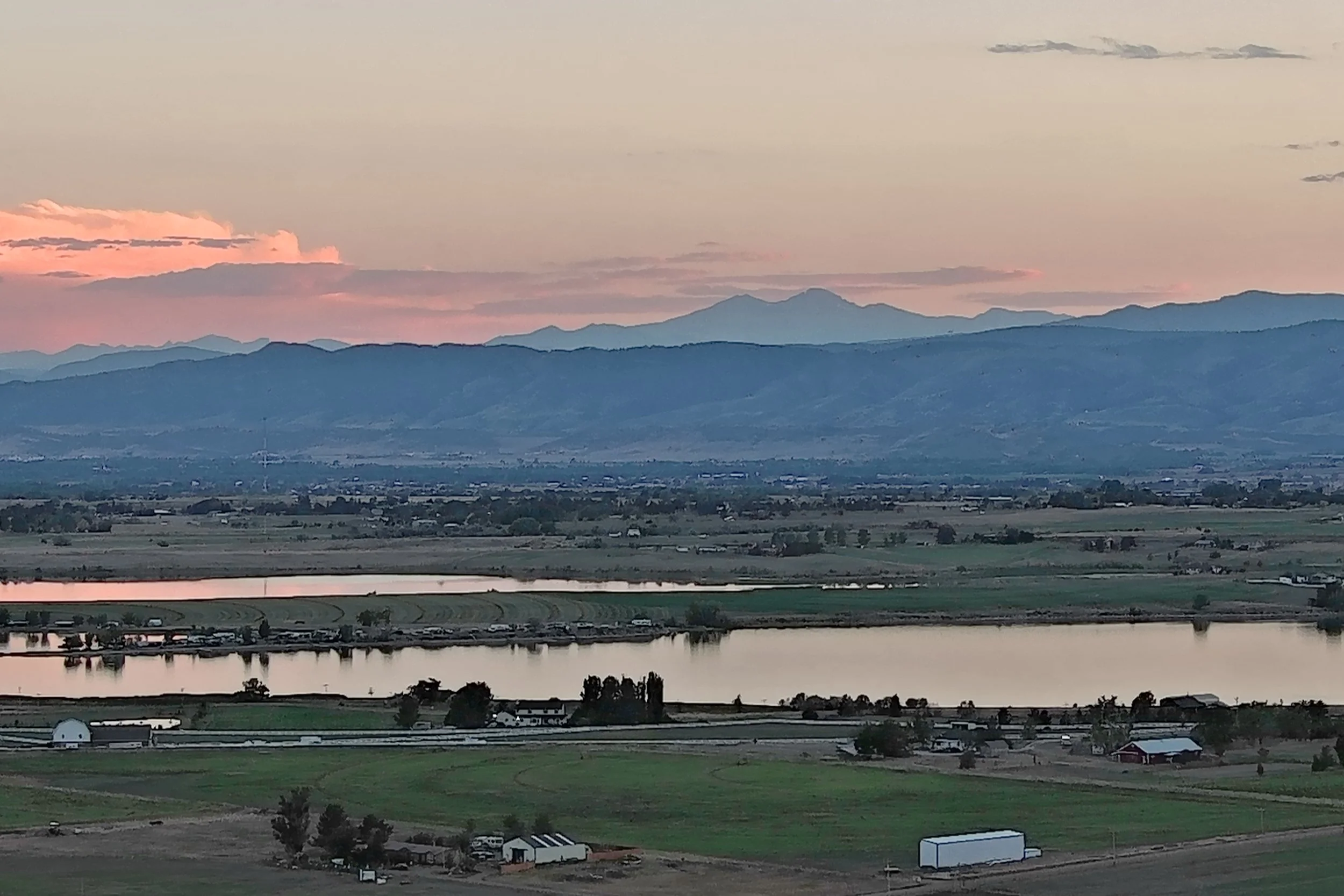 Scenic view of a mountain range during sunset with a body of water, fields, and farm structures in the foreground.