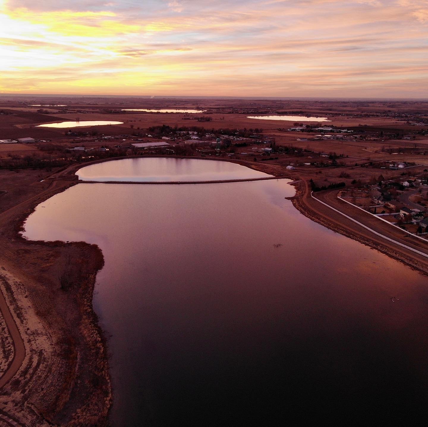 Aerial view of a lake at sunset with reflections of the colorful sky on the water, surrounded by a rural landscape with roads, fields, and scattered buildings.