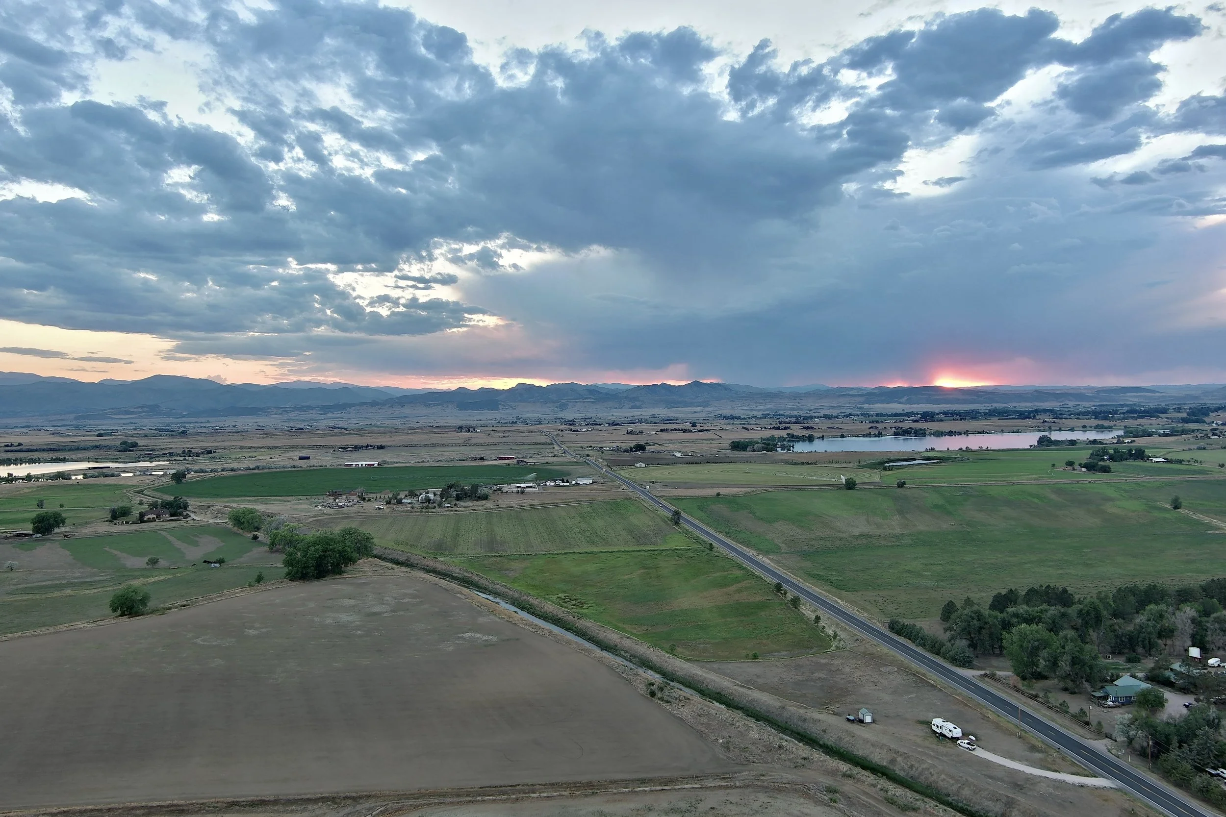 Scenic landscape with green farmland, a body of water, mountains in the distance, and a partly cloudy sky at sunset.