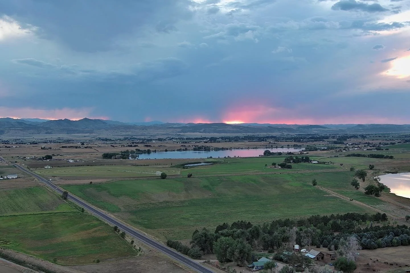 Scenic view of farmland with green fields and ponds, a highway runs through the area, mountains in the distance, stormy clouds overhead, and a pink sunset glow on the horizon.