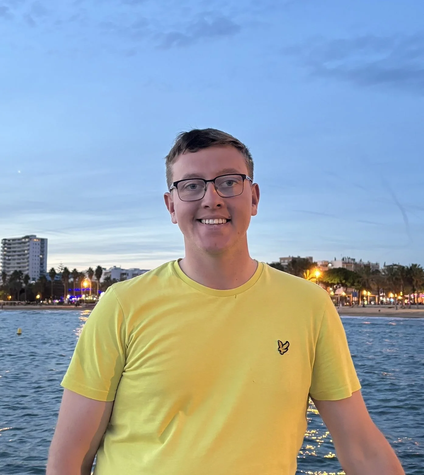 A young man with glasses wearing a yellow T-shirt stands outdoors at sunset with a city skyline, water, and illuminated buildings and trees in the background.