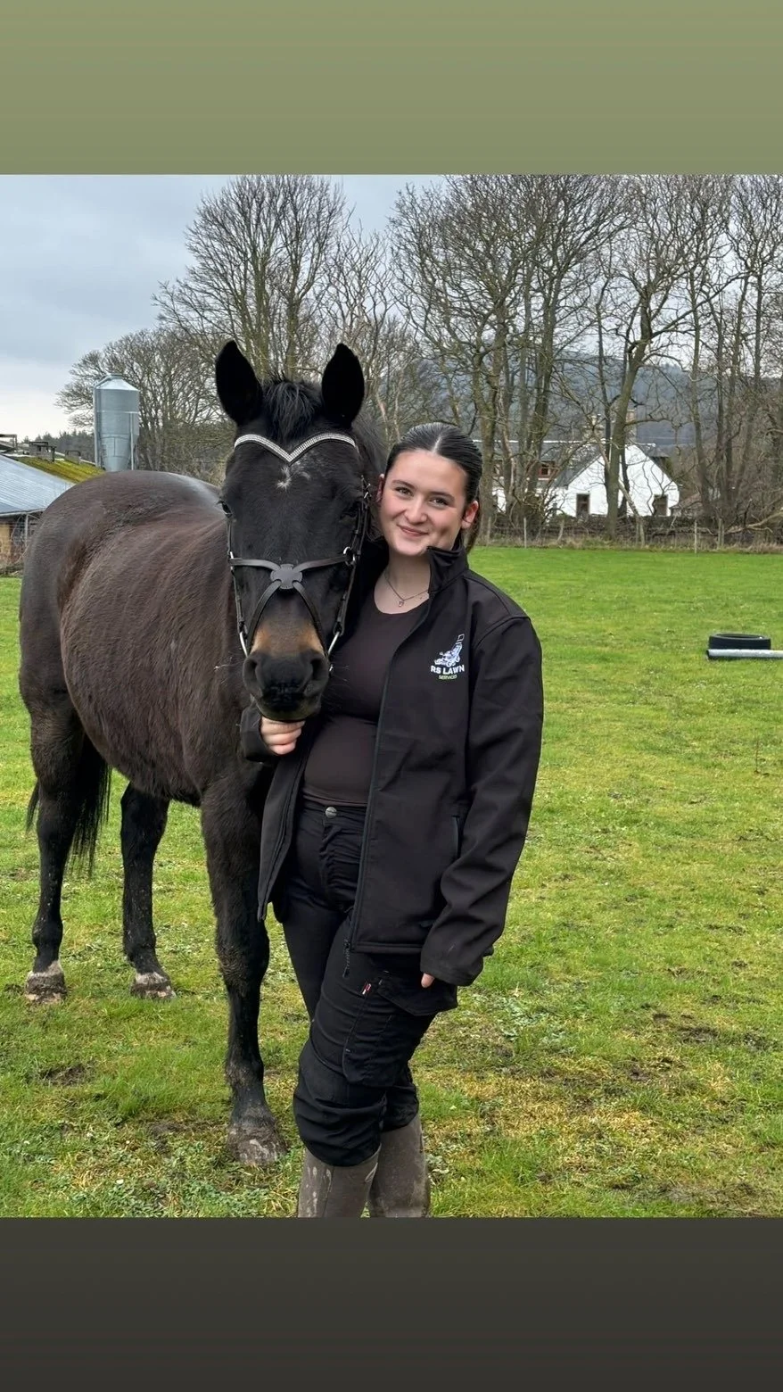 A young woman standing next to a dark brown horse with a black mane outdoors on a grassy field, with leafless trees and farm buildings in the background.