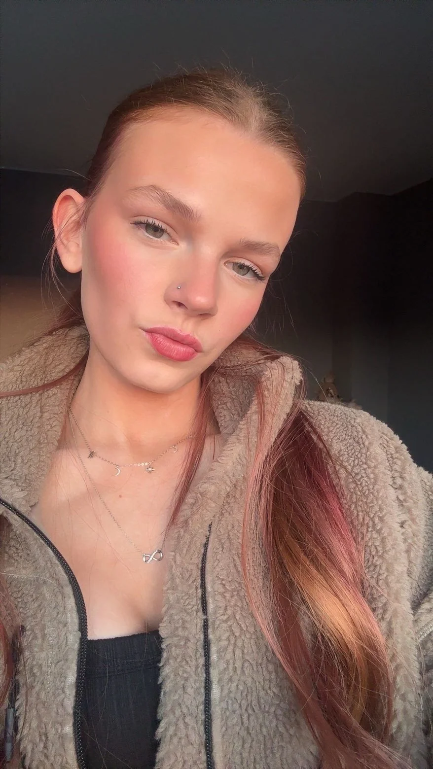Close-up image of a young woman with red hair, wearing layered necklaces and a beige fleece jacket, indoors with dark background.