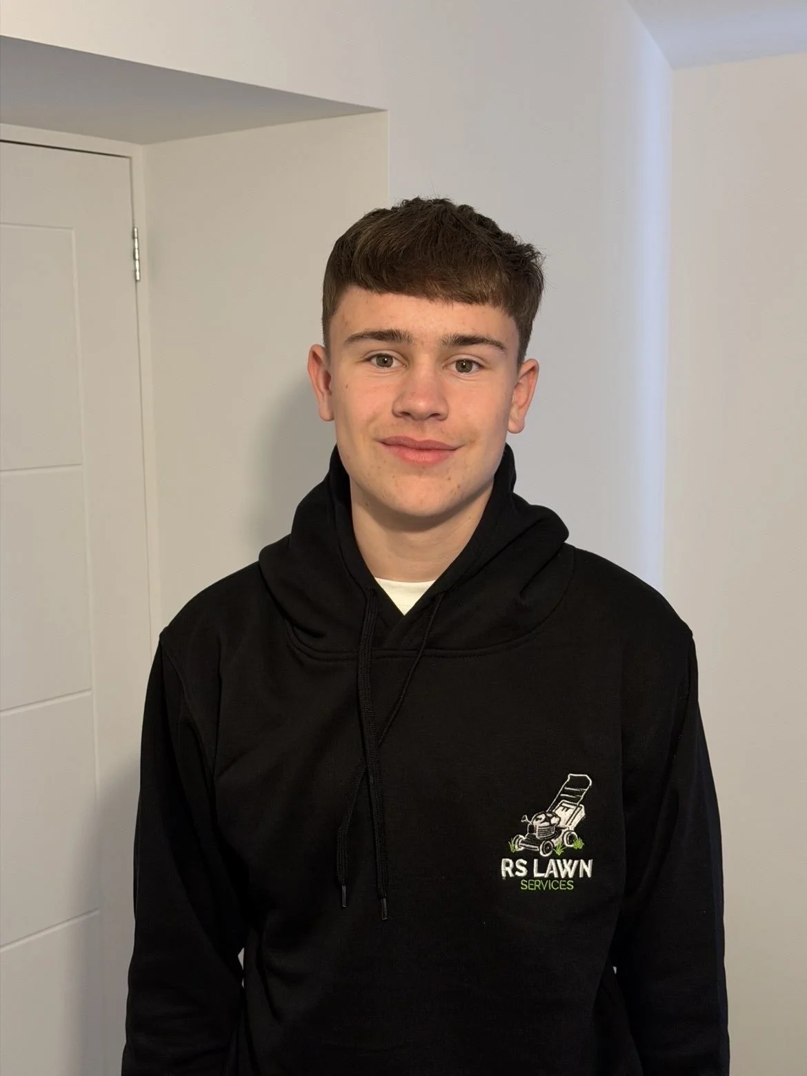 A young man with short brown hair and light skin, wearing a black hoodie with a logo for R S Lawn Services, standing indoors against a plain white wall.