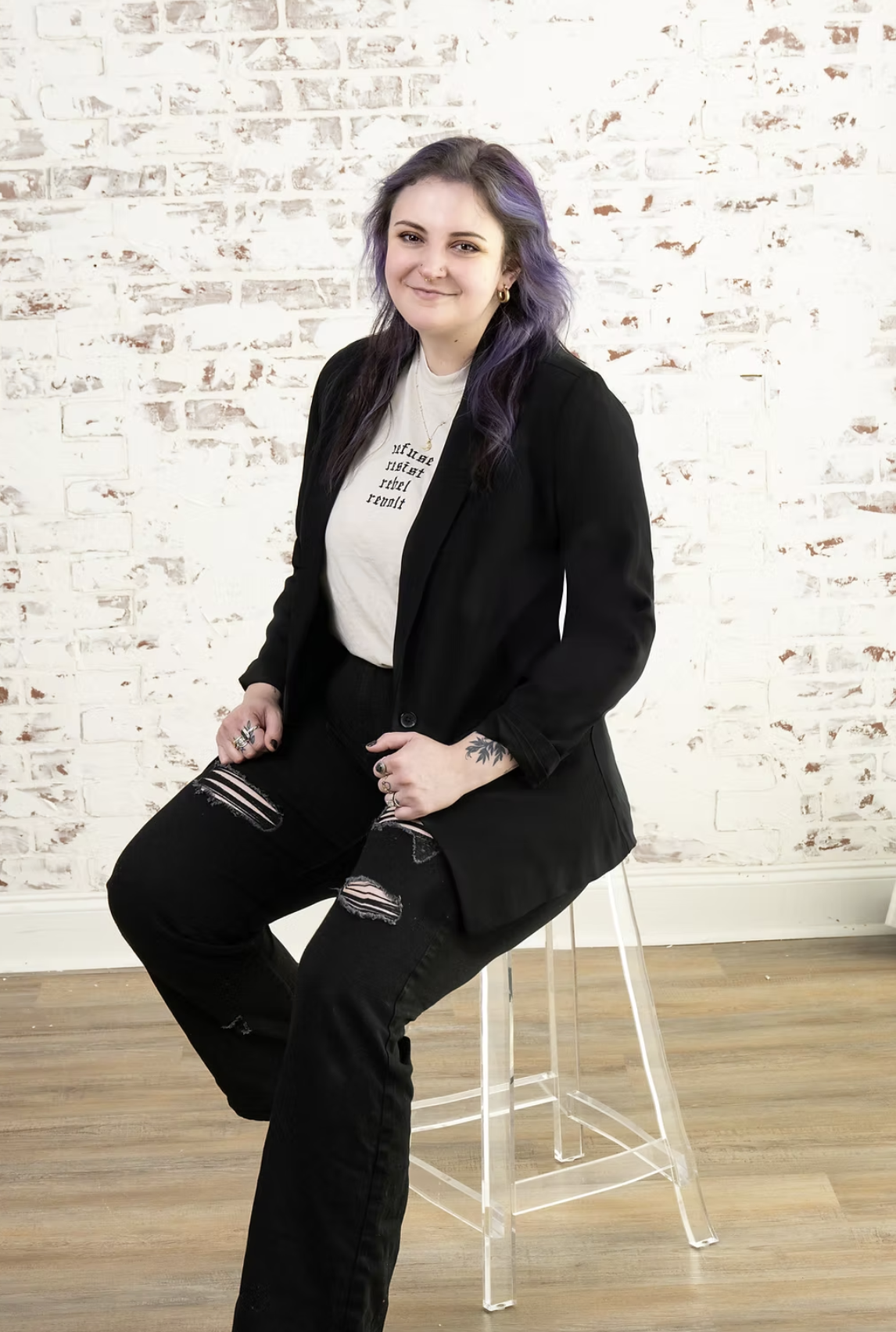 A young woman with purple hair sitting on a clear acrylic chair in front of a white brick wall, wearing a black blazer, ripped black jeans, and a white T-shirt.