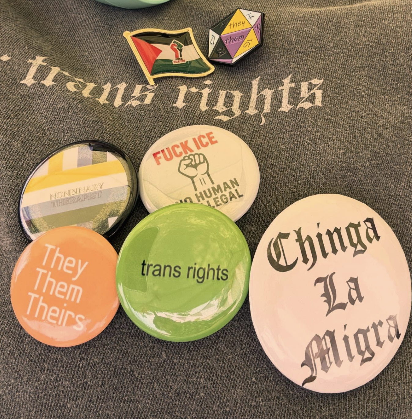 A collection of buttons and pins with various messages and symbols on a gray fabric surface. The buttons include messages like 'trans rights,' 'fuck ICE and no human is illegal,' 'they/them/theirs,' and 'nonbinary therapist.' There are also stickers with political and cultural messages, including a Palestinian flag, and a die with gender pronouns.