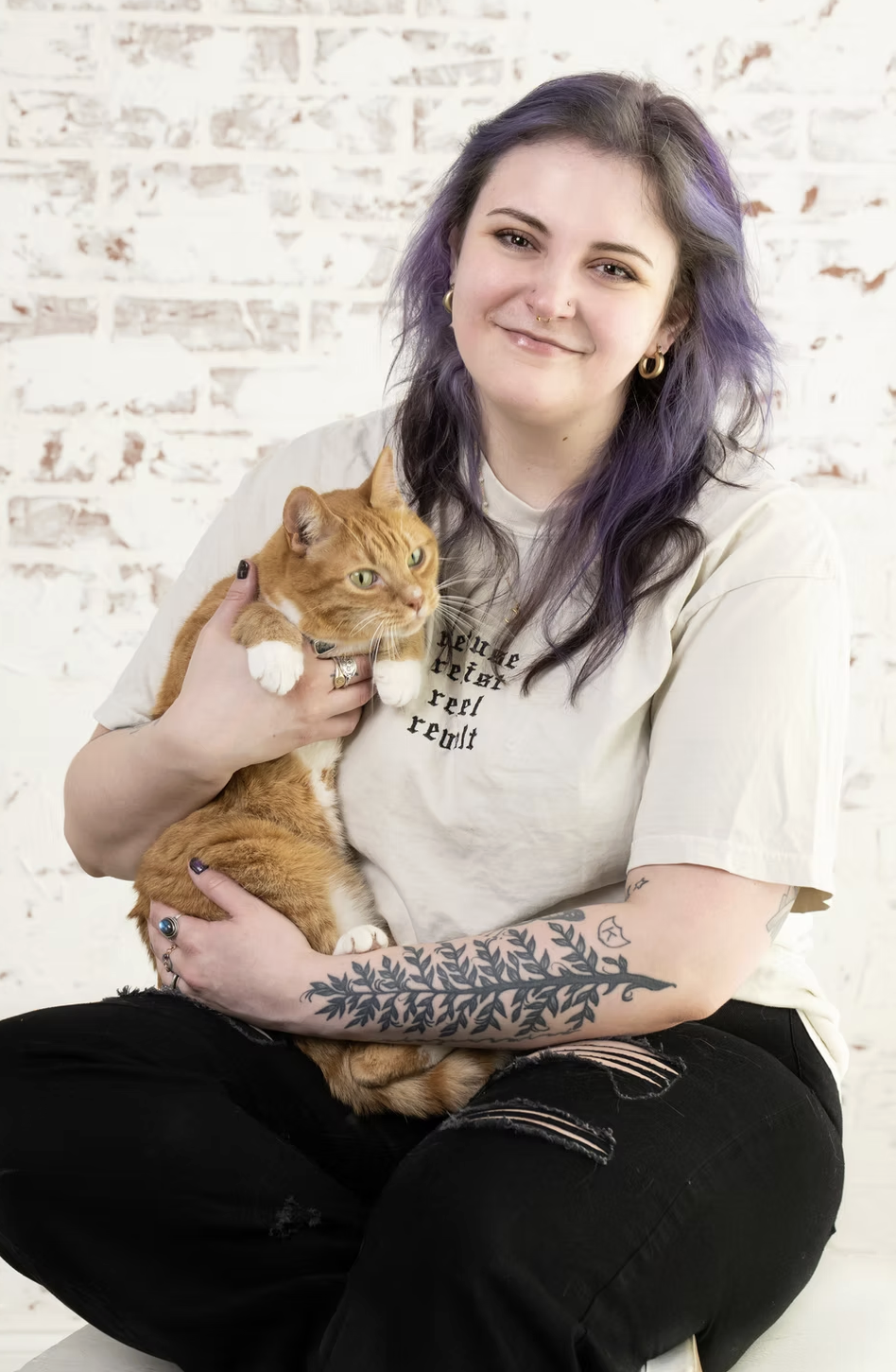 A smiling young woman with purple hair, wearing a beige t-shirt, holds an orange tabby cat. The woman has tattoos on her arm, piercings, and is sitting in front of a white brick wall.
