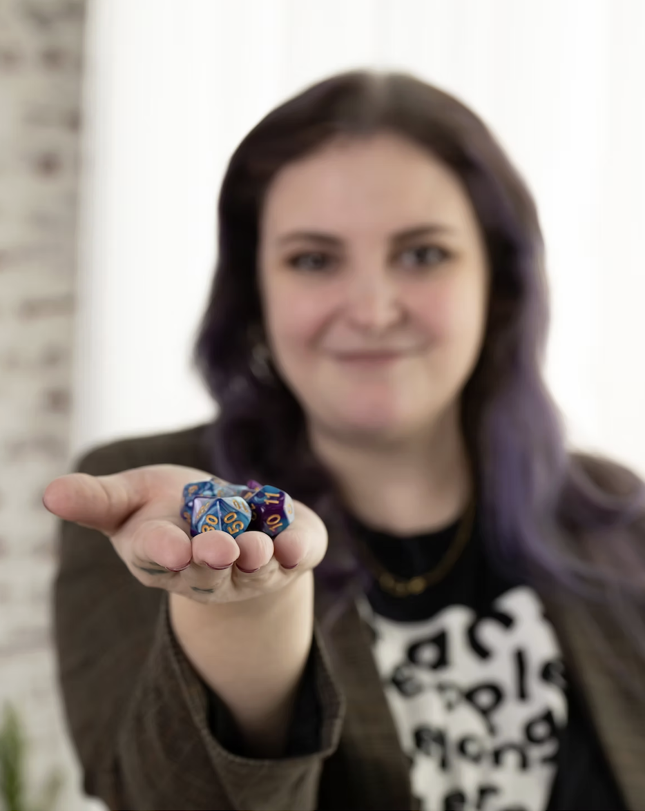 Woman holding several blue polyhedral dice with numbers visible, smiling, indoors with a blurred background.