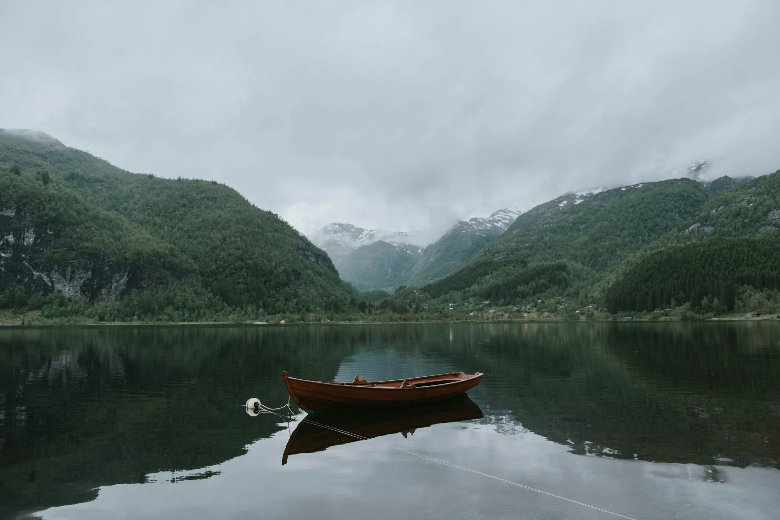 A small wooden boat floating on a calm lake with reflections of green mountains and cloudy sky, with snow-capped peaks in the distance.