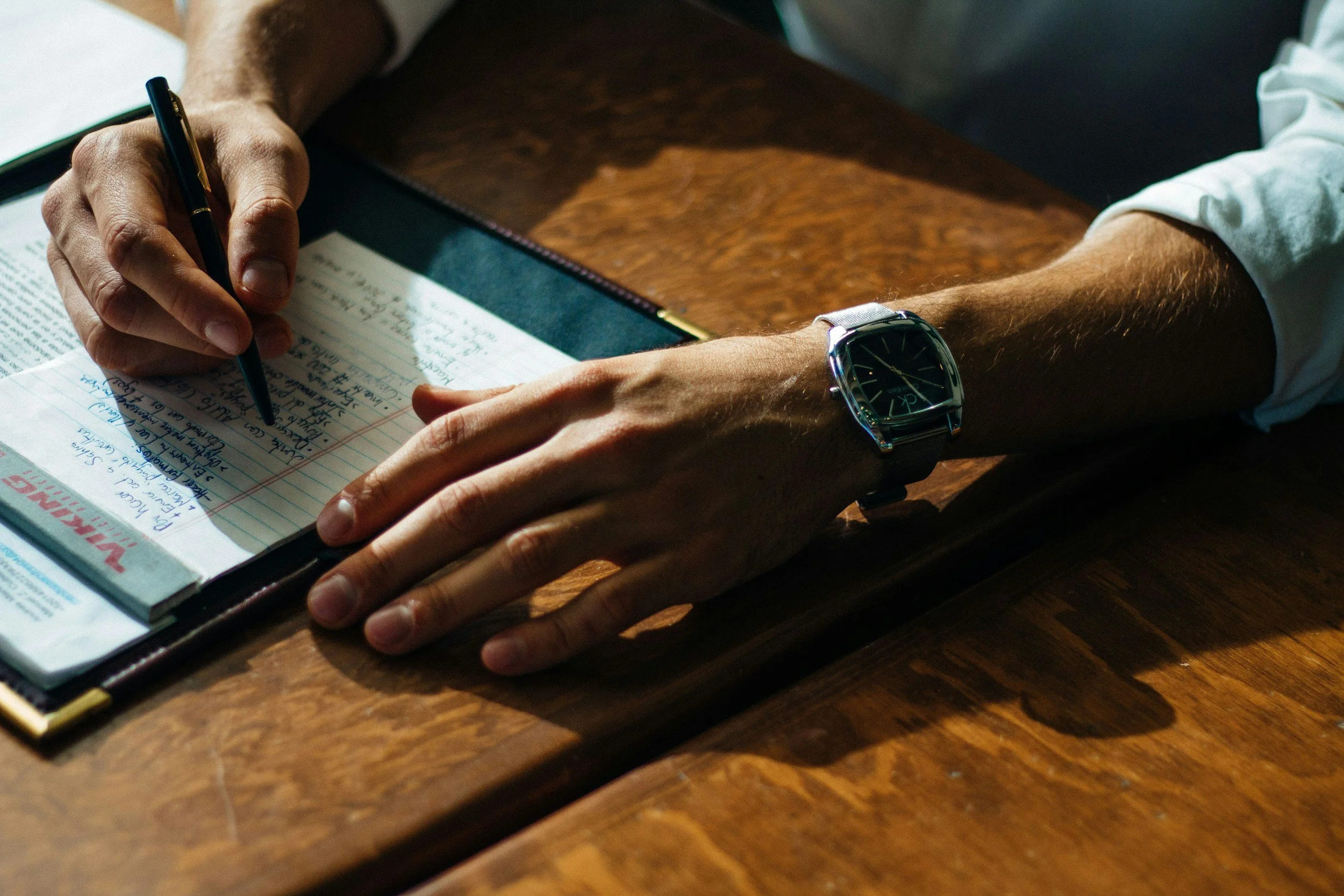 A person writing on a lined notepad placed on a wooden table. The person's left hand is resting on the table, wearing a wristwatch, while the right hand is holding a black pen. The person's forearms are visible, with rolled-up sleeves.