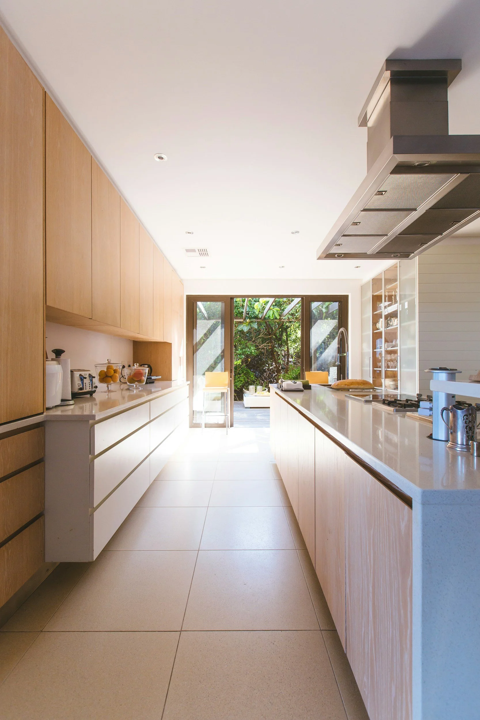 A modern kitchen with white countertops, light wooden cabinets, a stainless steel range hood, and a large window overlooking a garden.