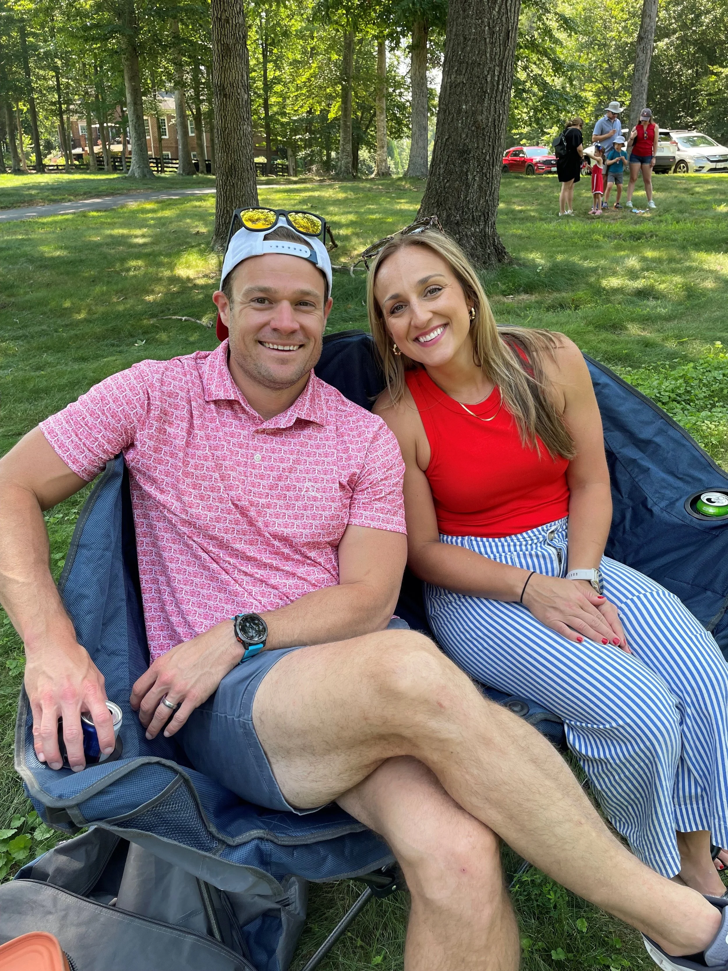 Smiling man and woman sitting on a blanket in a park with trees and other people in the background.