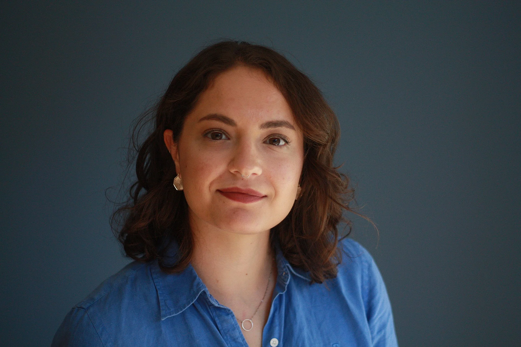 A woman with brown hair wearing a blue shirt and silver earrings against a dark background