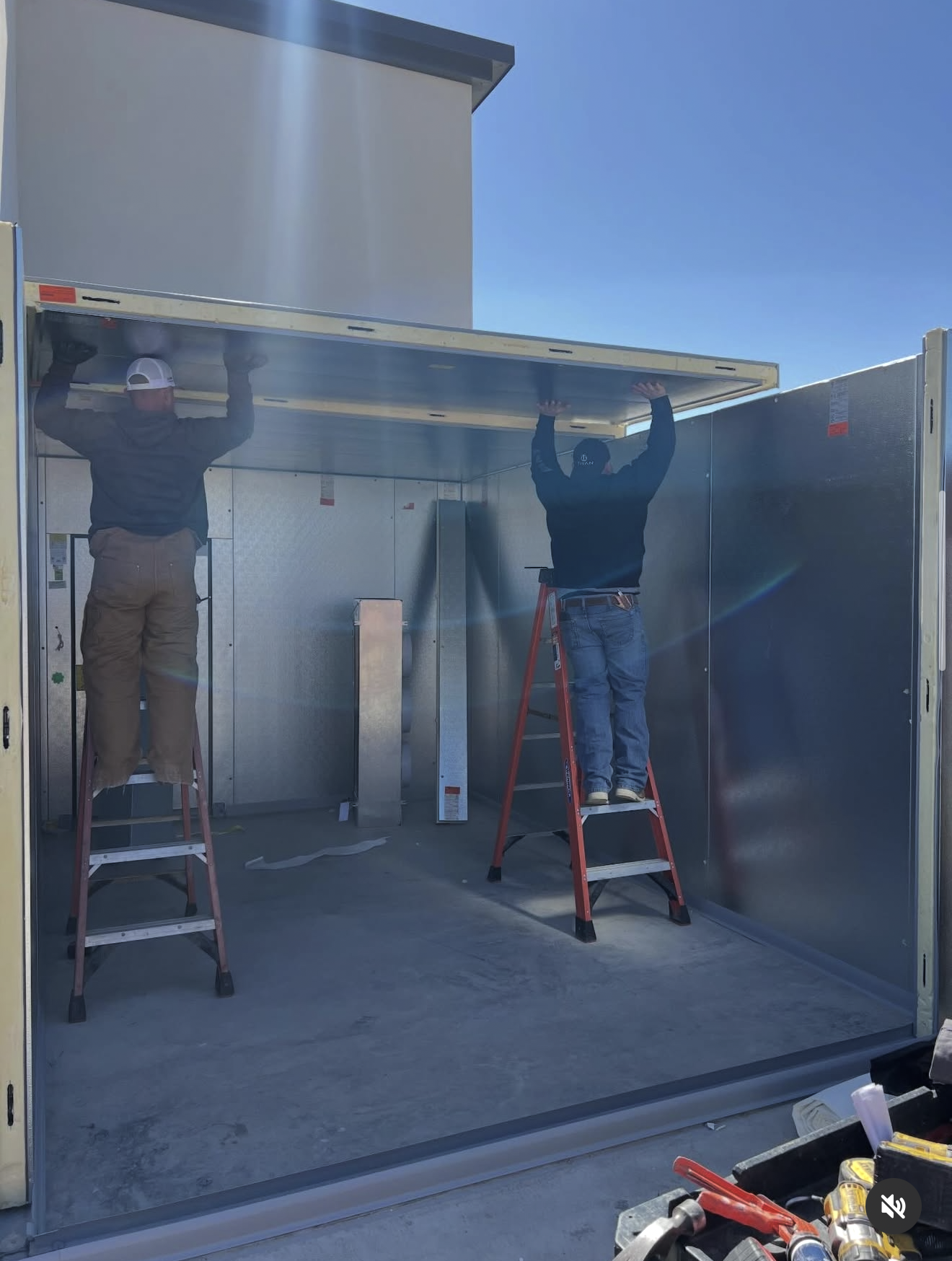 Two workers on ladders are installing or repairing a large, rectangular metal structure or cabinet outdoors under clear blue sky.
