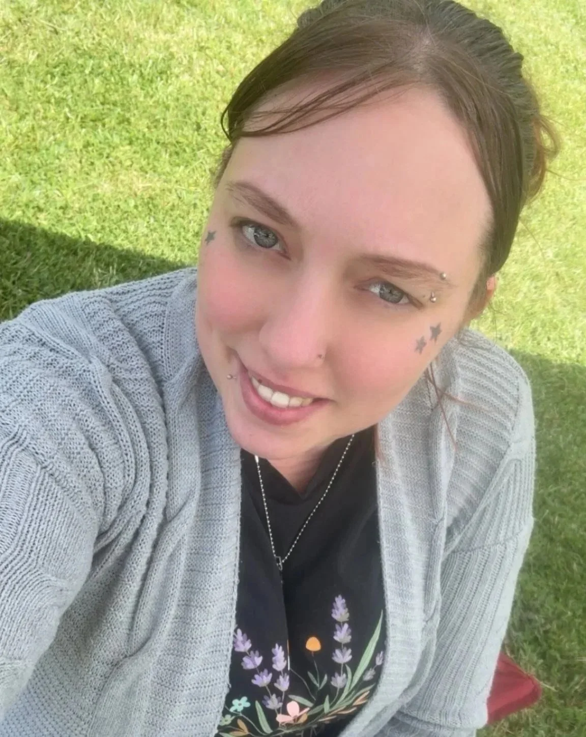 A young woman with dark brown hair, tattoos on her face including stars, is smiling and taking a selfie outdoors on a sunny day, with green grass in the background.