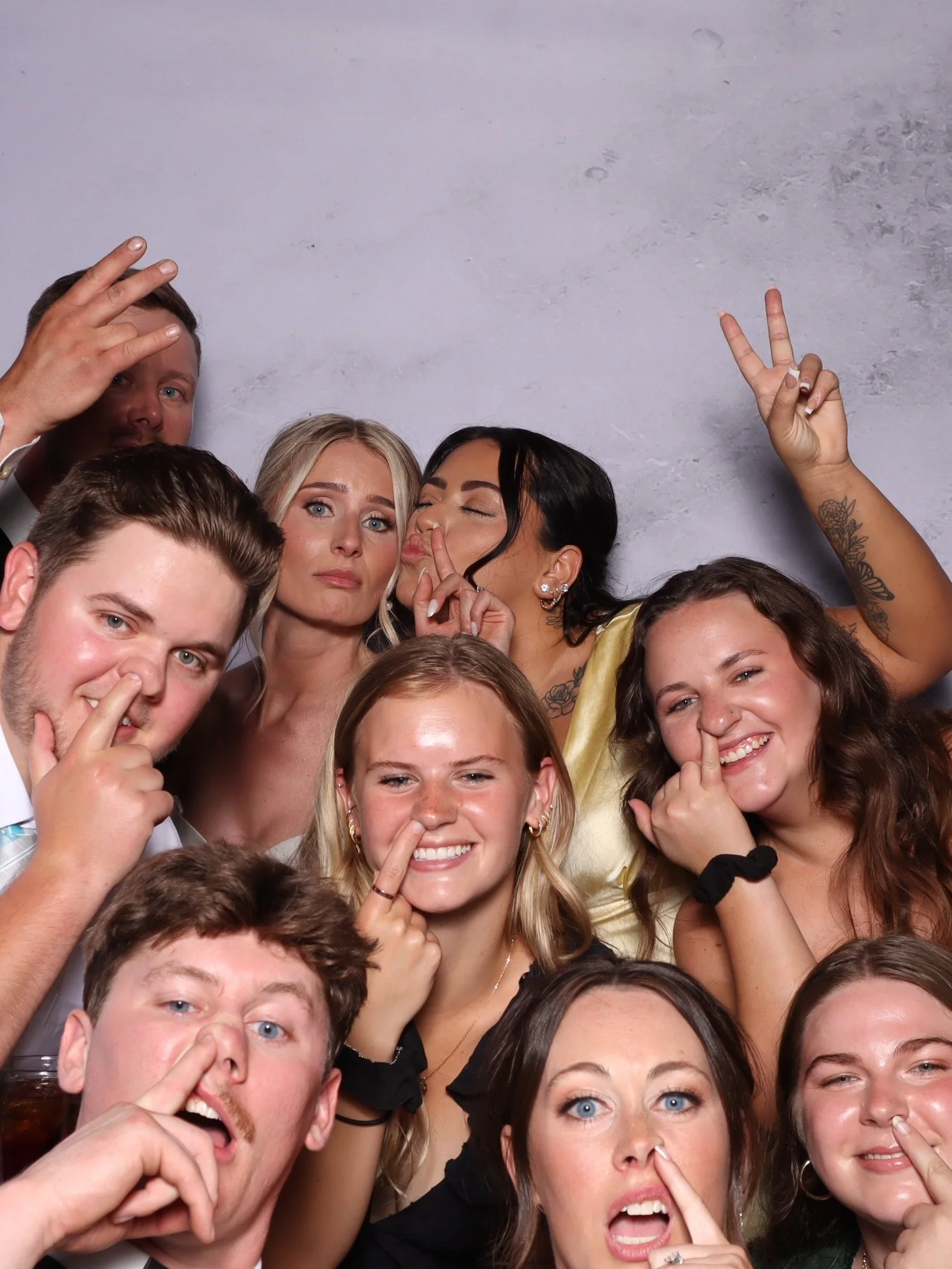 A group of diverse young adults gathered closely together, making playful gestures such as peace signs and poking their noses, against a plain gray background. They are smiling, laughing, and appear to be enjoying themselves at a social event or cele