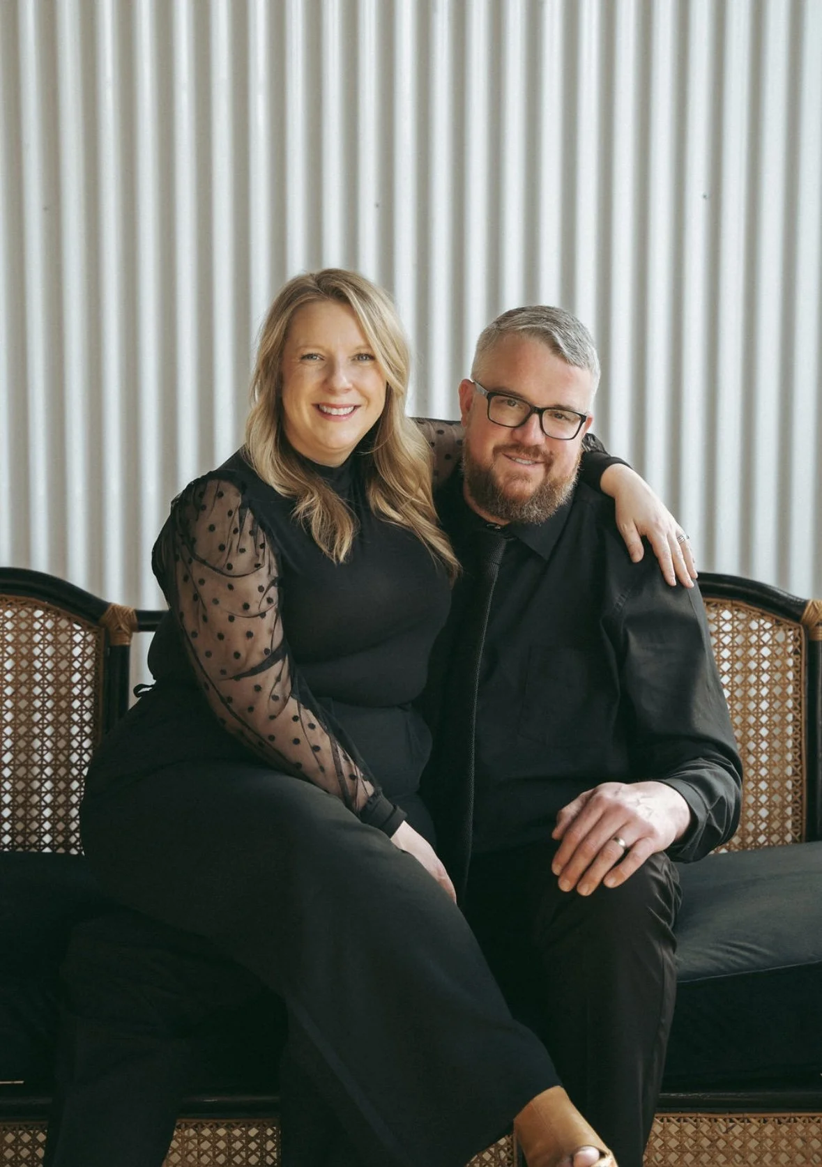 A smiling woman with blonde hair and a man with gray hair and glasses sitting together on a bench, in front of a light-colored striped curtain background.