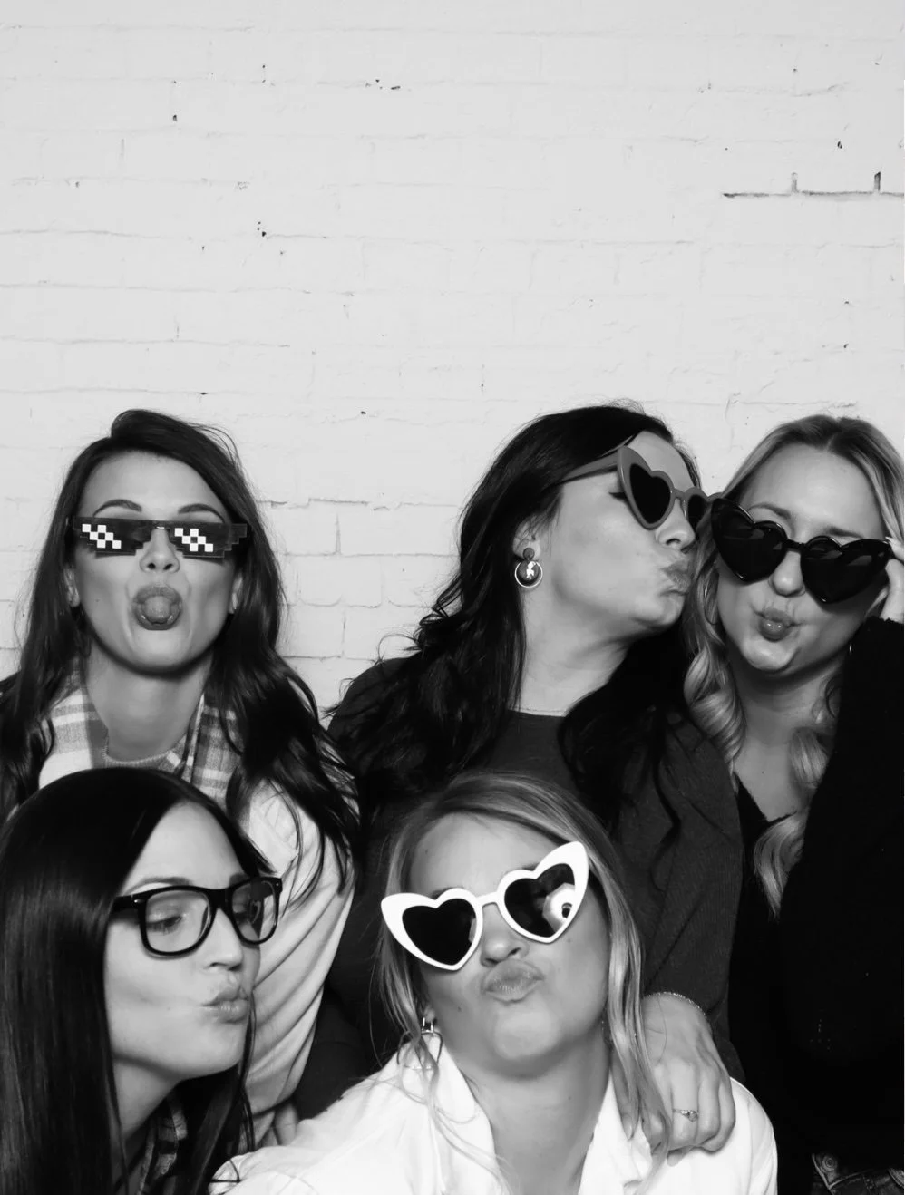 Group of five women posing with funny sunglasses and making puckered lips against a white brick wall.