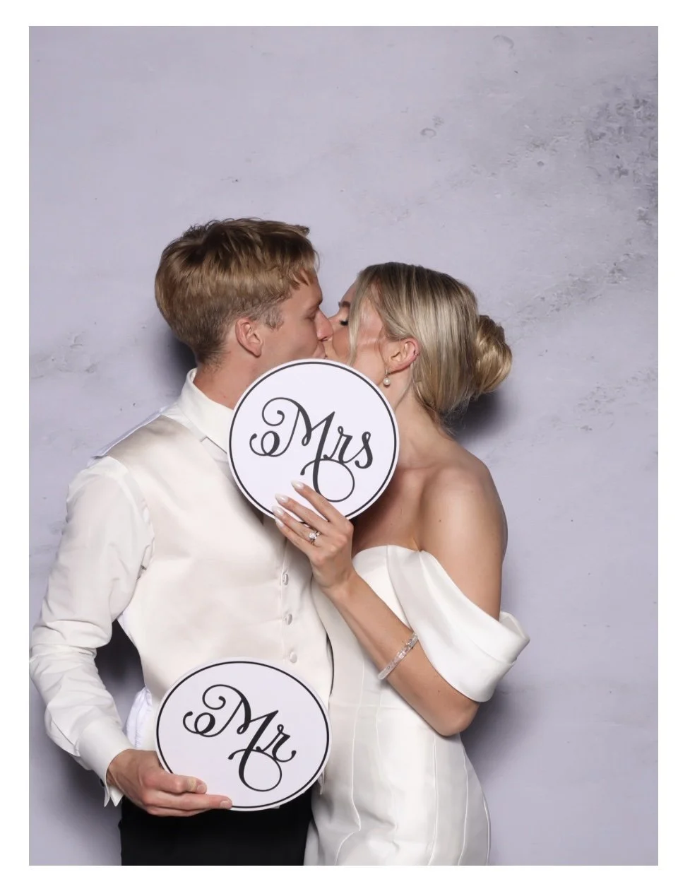 A bride and groom kiss while holding circular signs that say 'Mrs' and 'Mr' against a light gray wall.