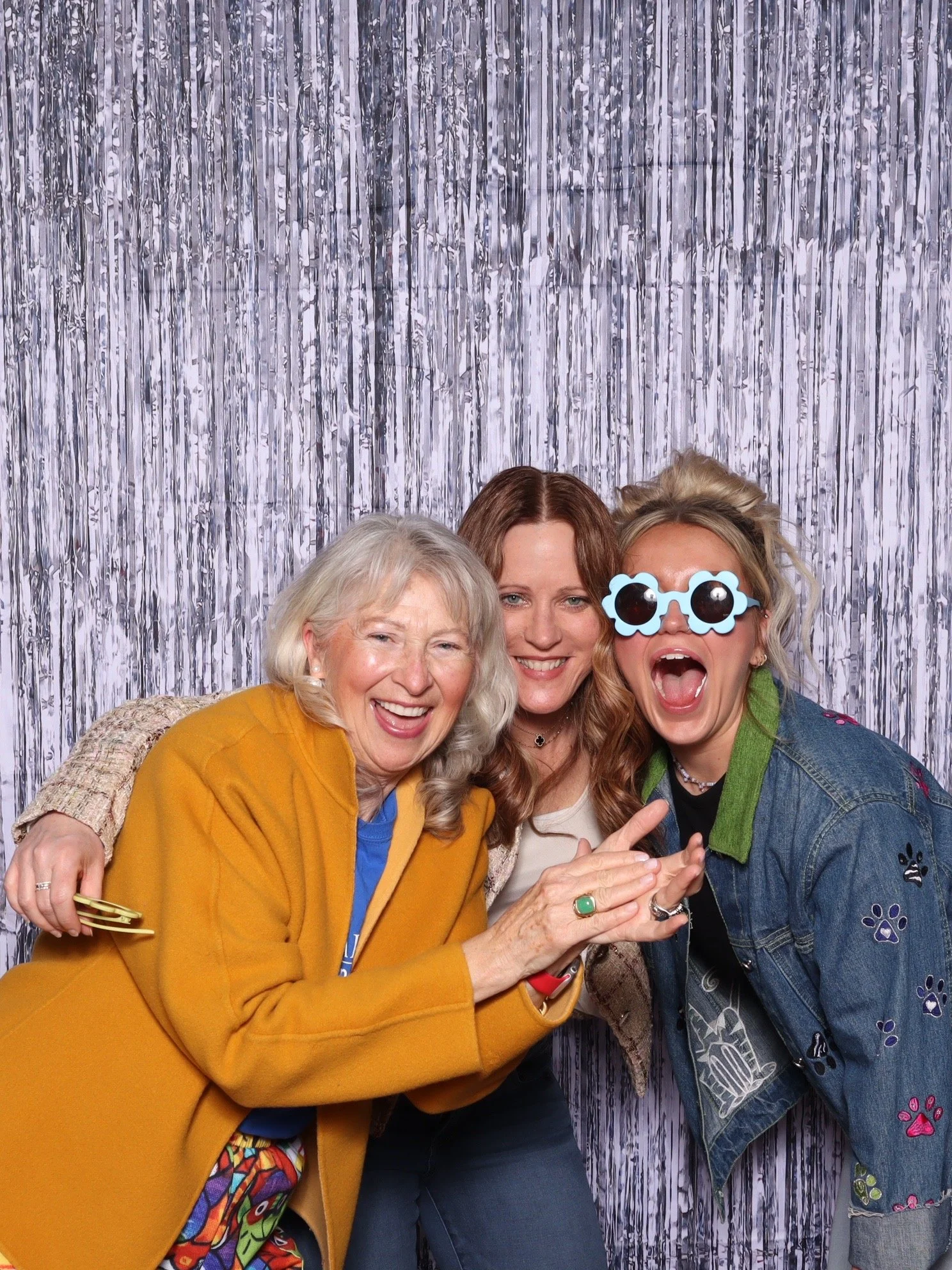 Four women of different ages posing together in front of a silver curtain backdrop at a celebration or photo booth, smiling and making joyful gestures.
