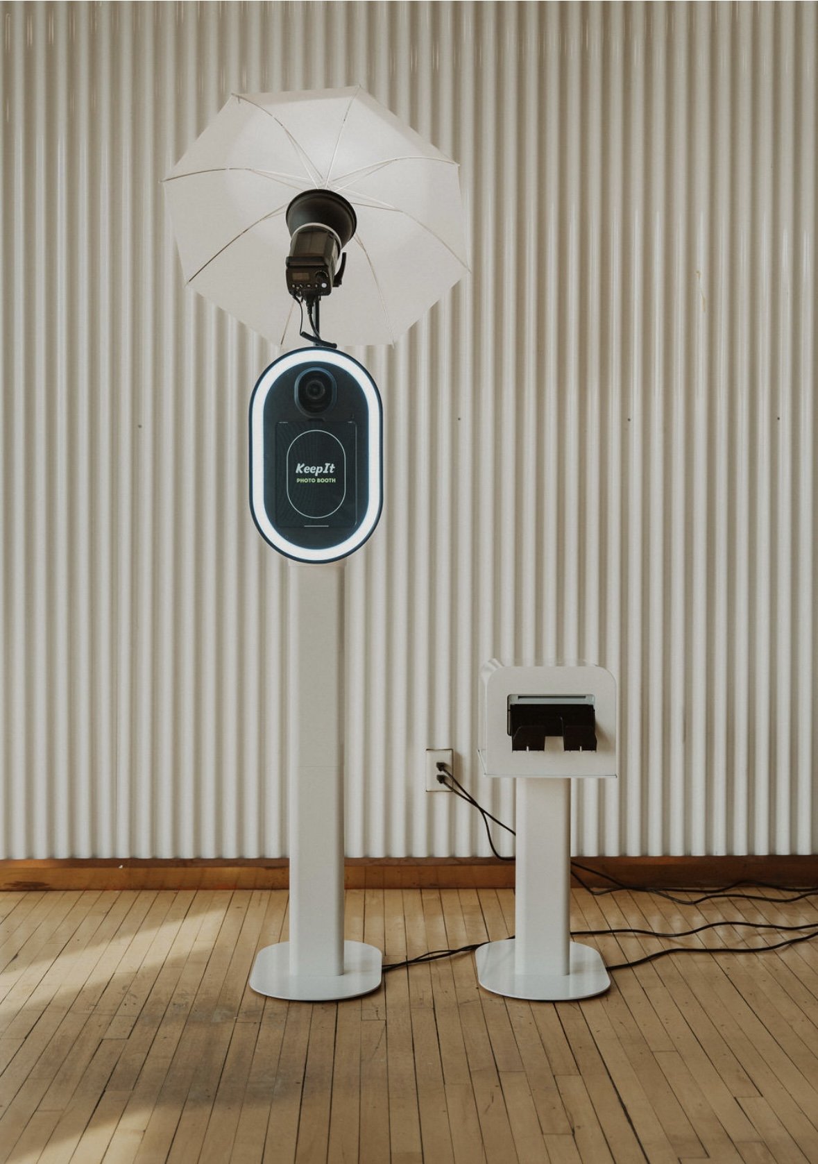 Photo booth with a clear plastic umbrella above and a vertical screen labeled "KeepIt PHOTO BOOTH" on a stand in front of a beige vertically ridged wall and wooden floor.