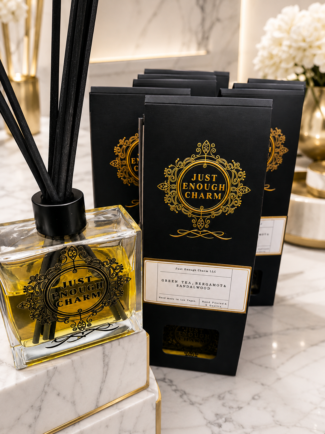 A display of black boxes labeled "Just Enough Charm" with gold decorative designs, along with a clear perfume bottle with similar gold branding, containing yellow liquid, on a white marble surface. In the background, there are gold and white vases with flowers.
