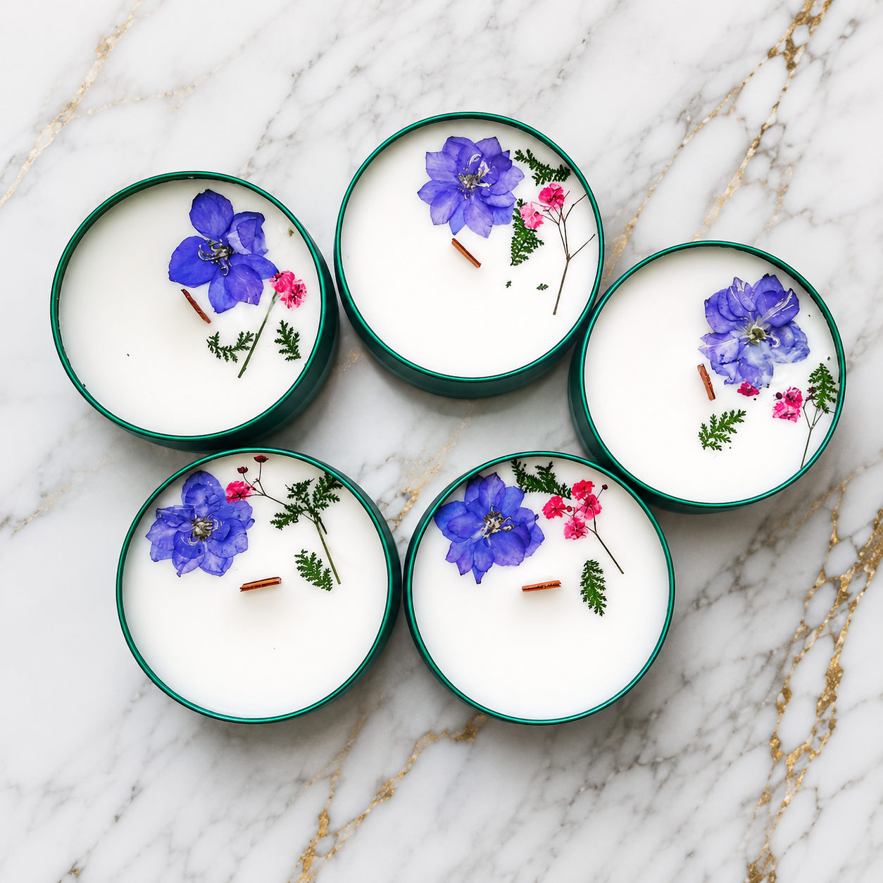 Five green containers of white scented candles decorated with blue and pink flowers and small green leaves on a white marble surface.