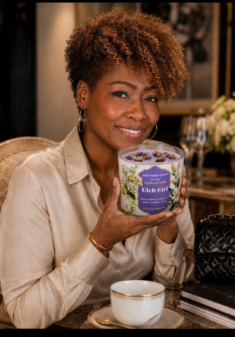 A woman with short curly hair, wearing a beige satin blouse and gold jewelry, smiling and holding a large scented candle labeled 'Rich Girl' at a table in a luxurious setting.