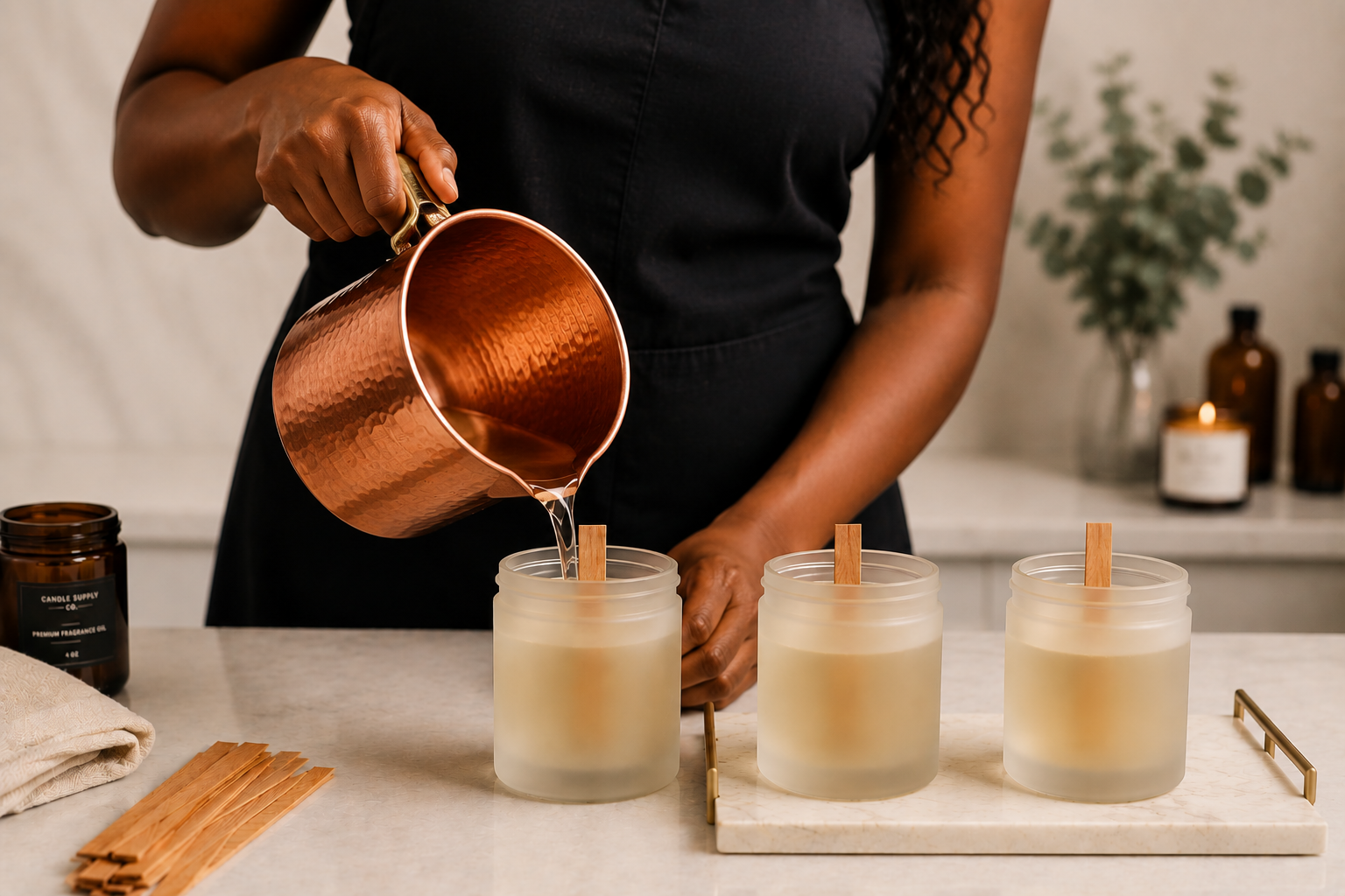Person pouring wax into three frosted candle jars on a marble tray, with candle wick sticks inside the jars, in a home setting with candles and plants in the background.