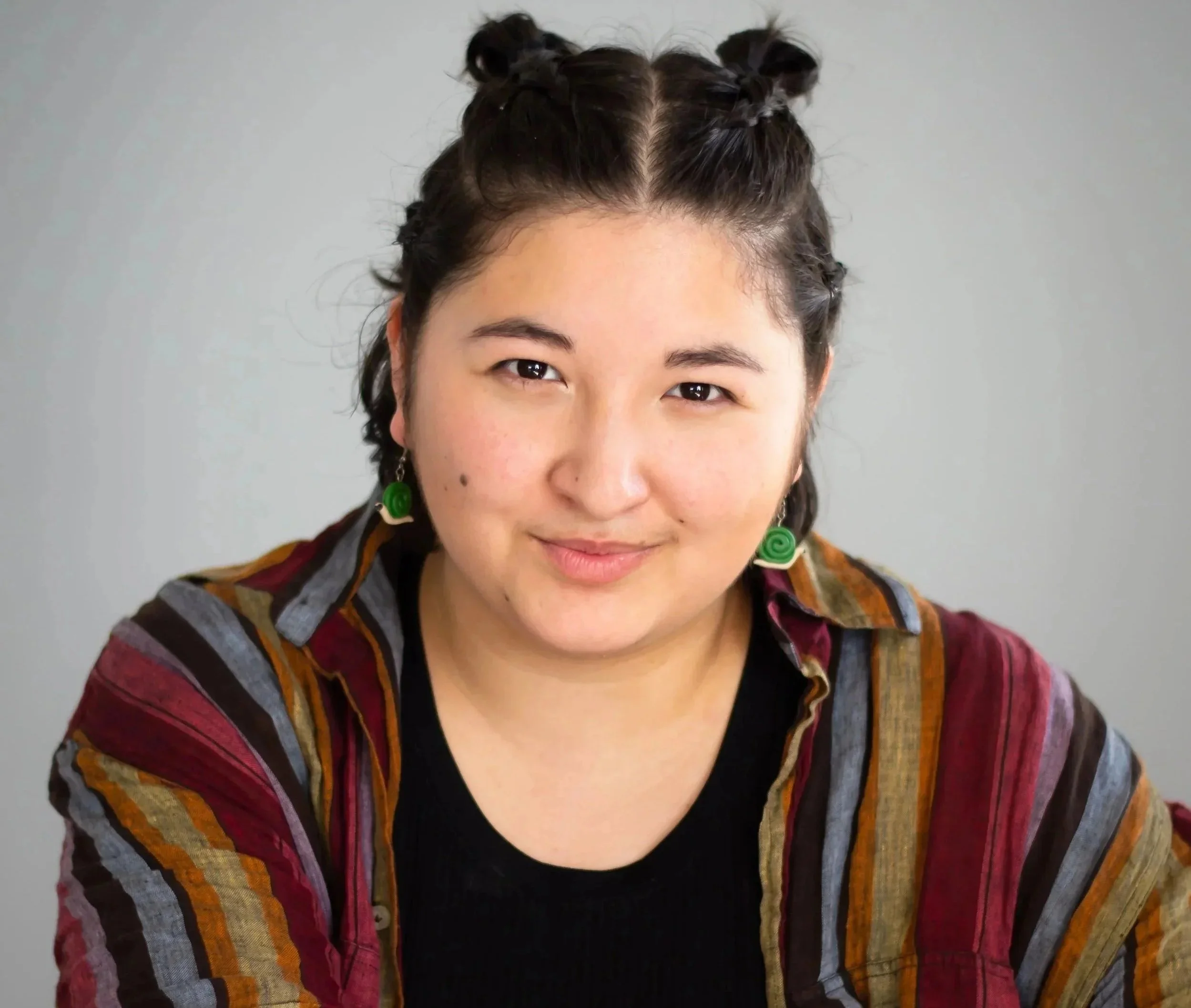 A asian theater and stage director with dark hair styled in two buns, wearing green spiral earrings and a colorful plaid shirt, smiling at the camera against a plain light gray background.